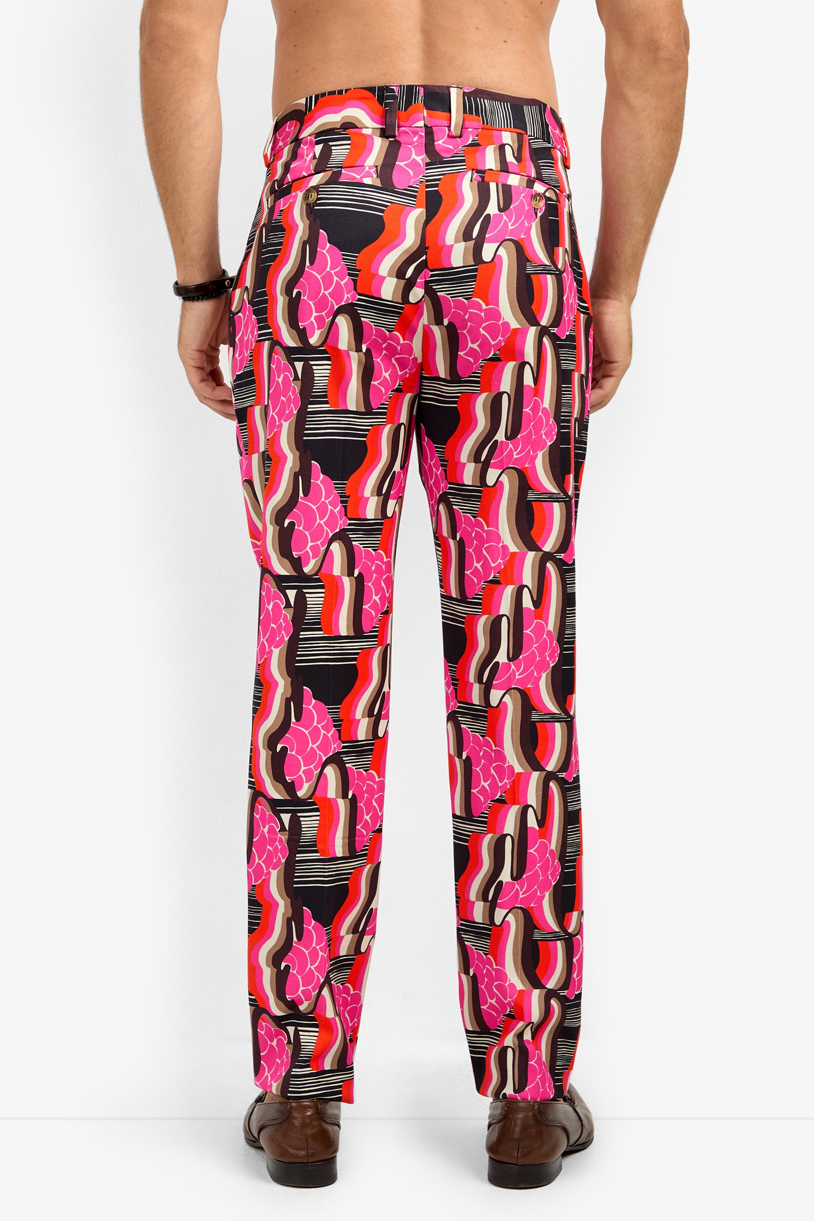 A man, shirtless and facing away from the camera, wears the CLYDE SLIM TROUSER featuring a bold abstract pattern in pink, red, black, and white, paired with brown shoes and a black bracelet.