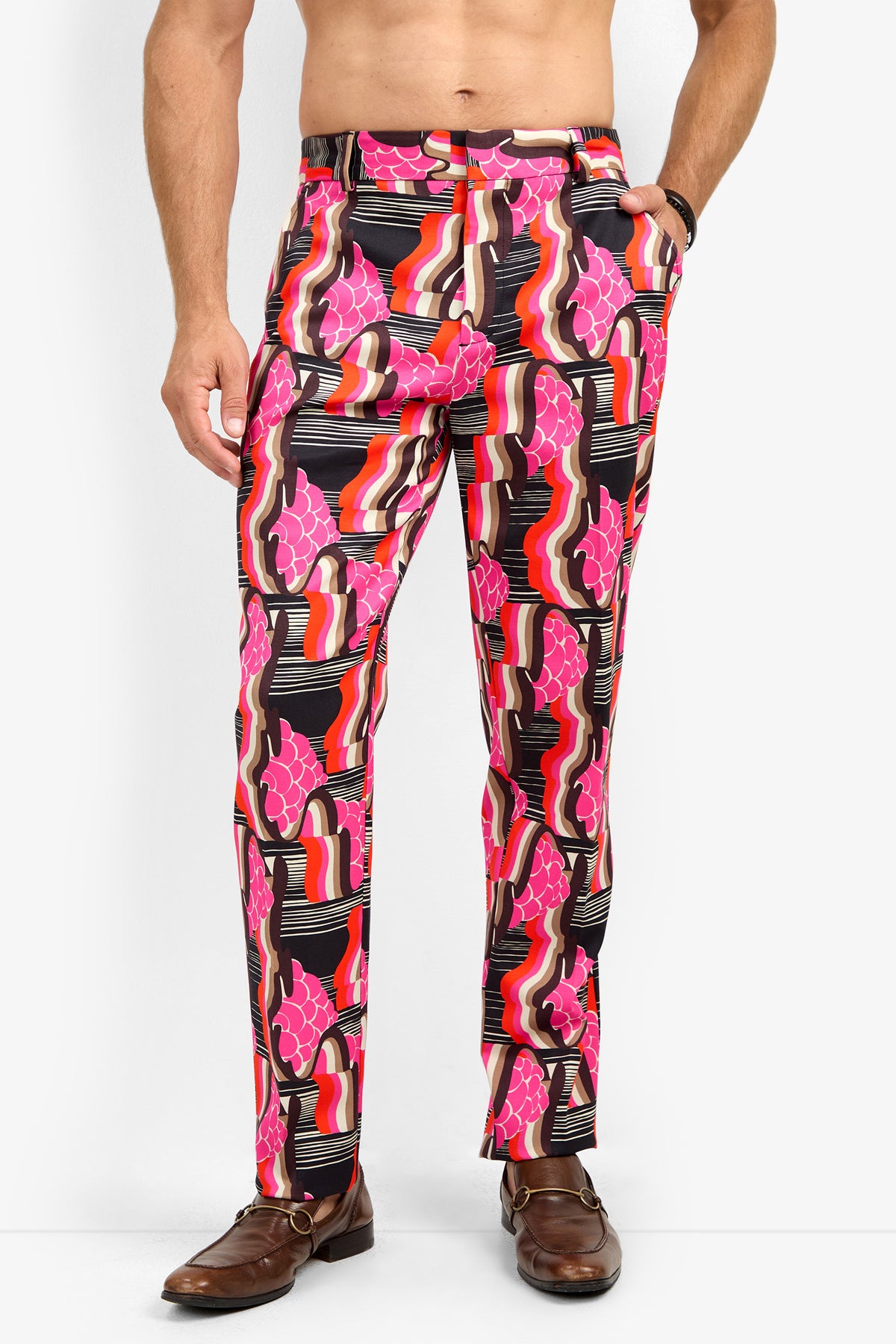 A man wears the CLYDE SLIM TROUSER, featuring colorful retro swirls in pink, red, black, and white. He pairs them with a brown belt and brown dress shoes. His upper body is bare, and his hands are tucked in his pockets.