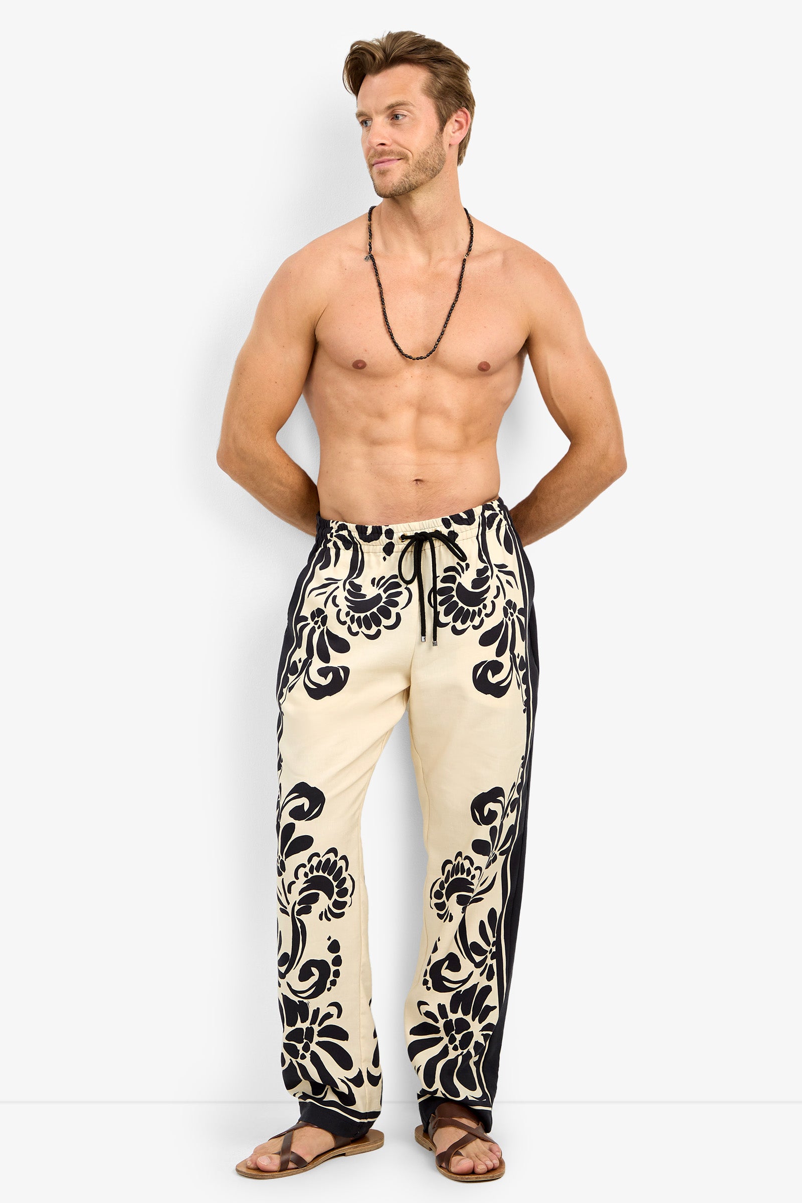 A shirtless man with a beaded necklace and sandals stands against a white background, hands in pockets, wearing the FELIPE DRAWSTRING PANT—cream-colored pants featuring bold black floral patterns.