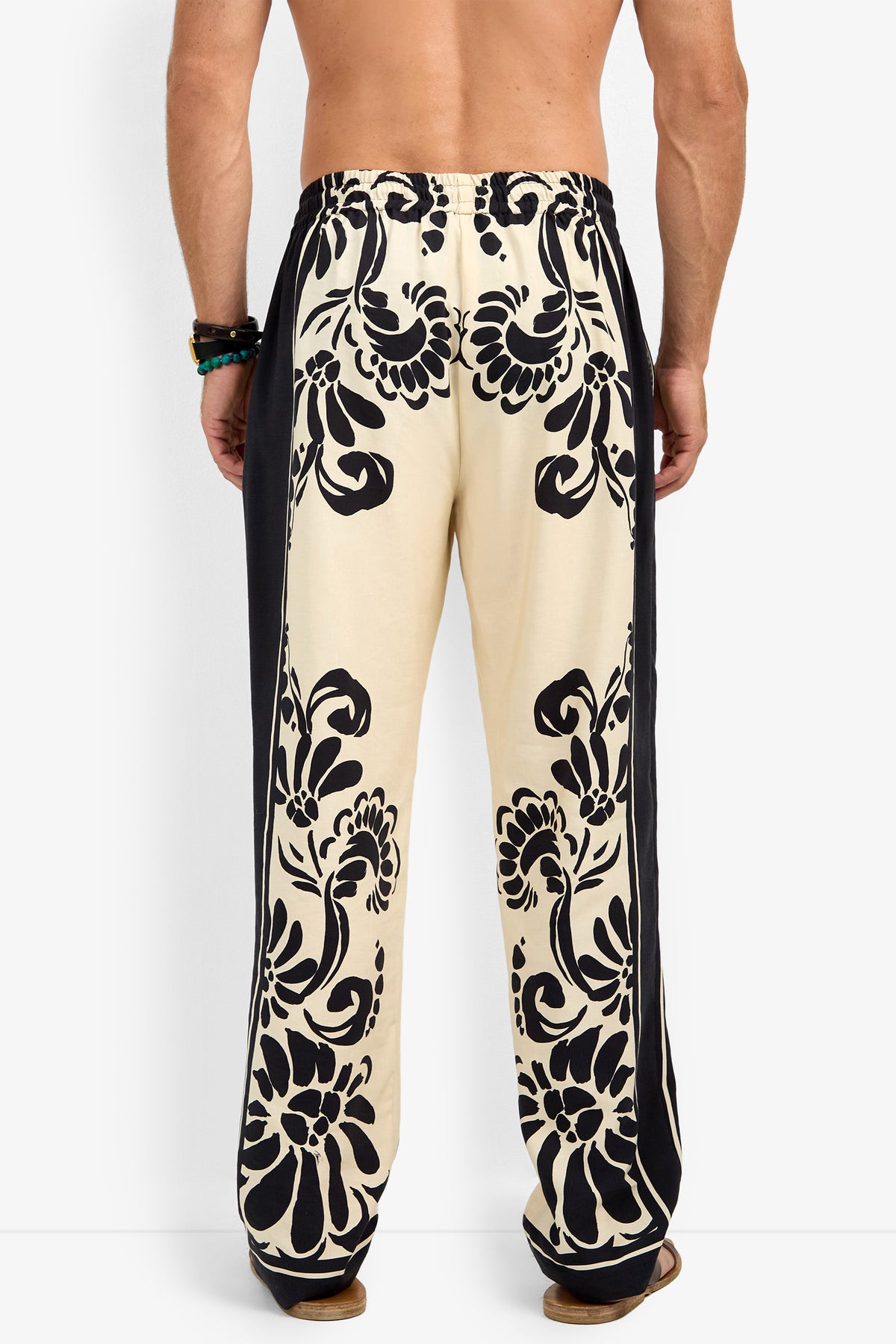 A shirtless man stands facing away, showcasing the back of the FELIPE DRAWSTRING PANT in cream with bold black floral patterns; his arms are relaxed by his sides and he wears beaded bracelets on one wrist.