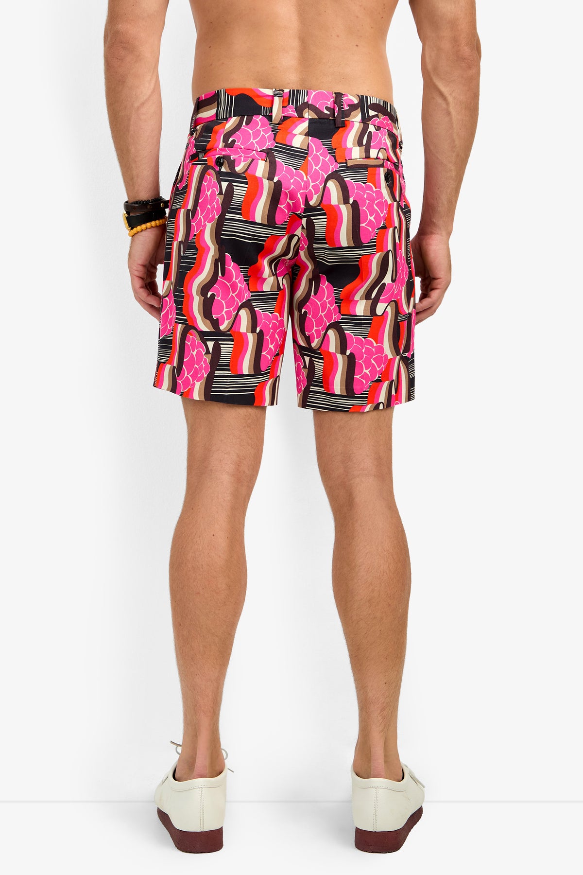 A person stands with their back to the camera in LAWRENCE SHORT—colorful, patterned shorts in pink, black, and orange—paired with white shoes and multiple bracelets on their right wrist against a plain white background.