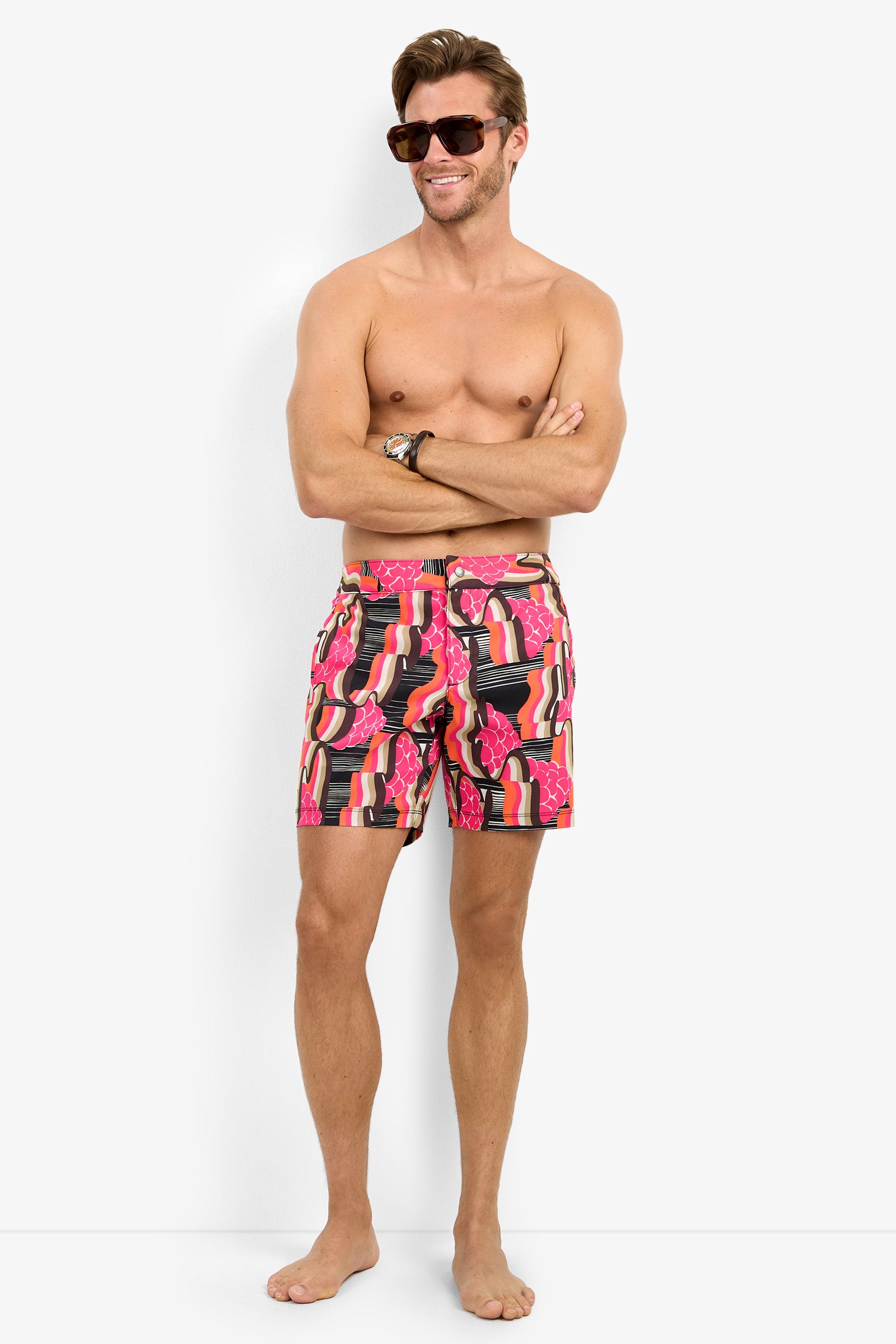 A smiling man stands barefoot against a white background, arms loosely crossed, wearing the HONOLULU BOARDSHORT and sunglasses.