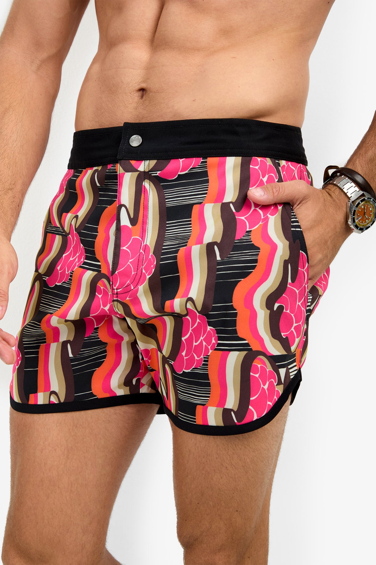 A person wears the SURFSIDE BOARDSHORT—retro-patterned swim shorts with black trim and waistband—standing against a white background, one hand in their pocket, wearing a watch.