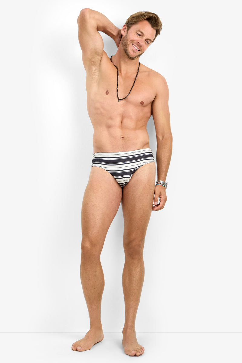 A man wearing the BONDI SWIM BRIEF and a necklace stands barefoot against a plain background, smiling with one arm raised to touch the back of his head.