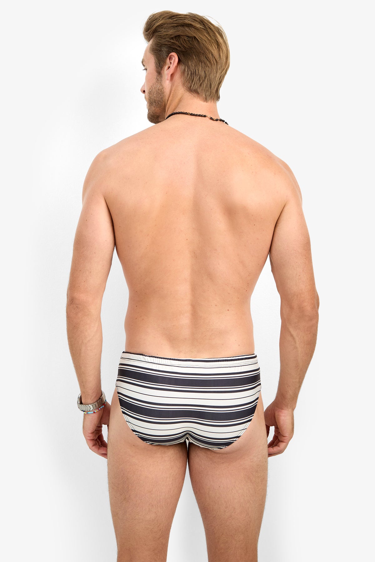 A man stands with his back to the camera, shirtless, wearing the BONDI SWIM BRIEF in black, white, and gray stripes. He accessorizes with a wristwatch and a black necklace.