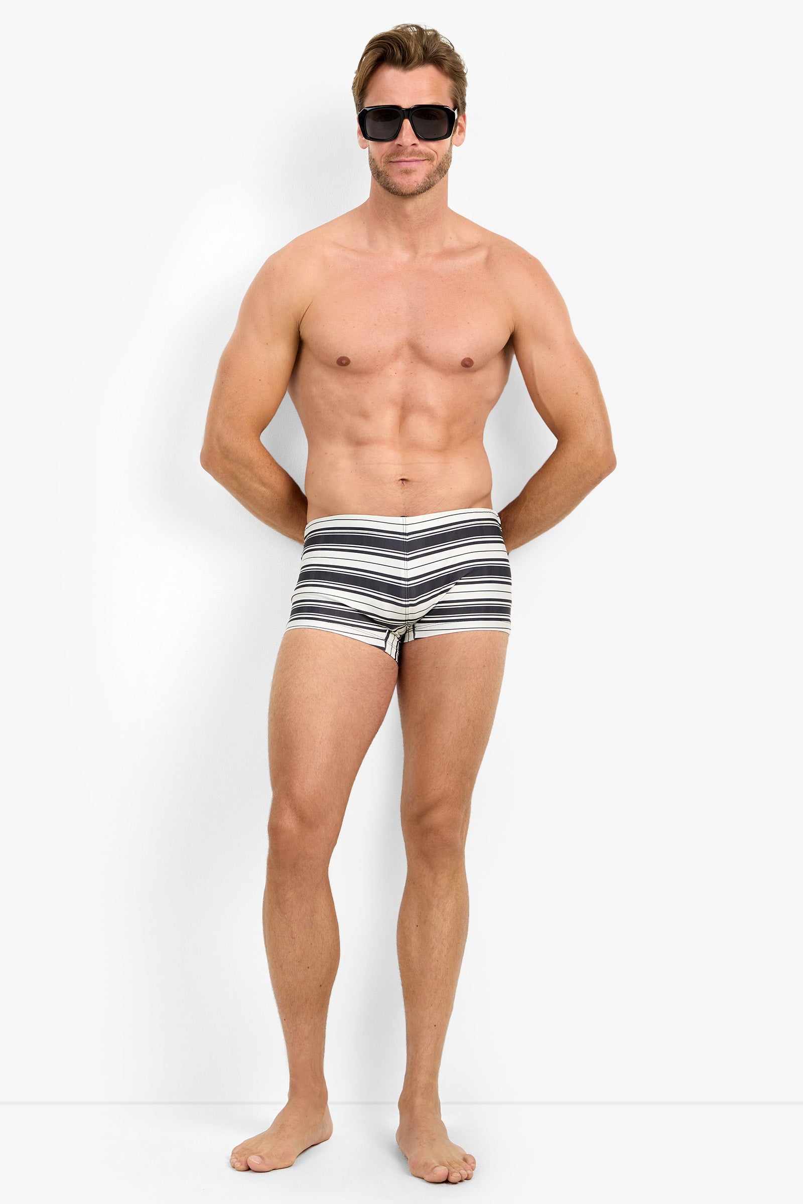 A shirtless man wearing sunglasses and MONTEGO BAY SWIM TRUNK stands barefoot against a plain white background, facing the camera with his arms relaxed at his sides.
