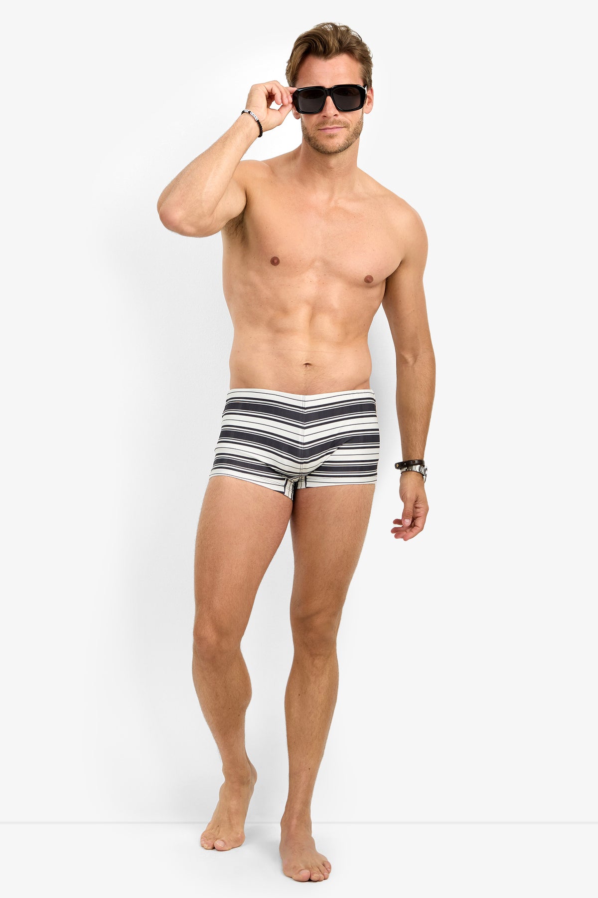 A shirtless man stands barefoot against a plain white background, wearing the MONTEGO BAY SWIM TRUNK in MULTI and sunglasses, holding the sunglasses with one hand and looking forward.