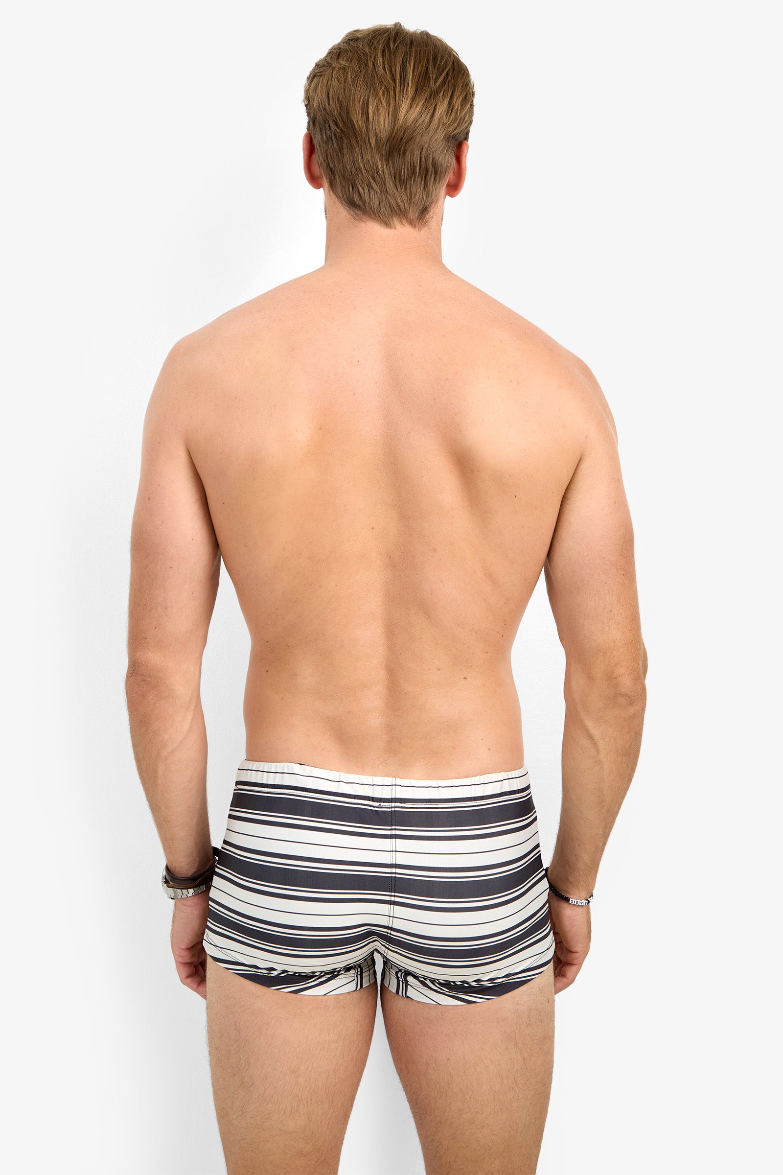 A shirtless man with light skin and short, light brown hair stands with his back to the camera, wearing MONTEGO BAY SWIM TRUNK in black, white, and gray stripes against a white background.