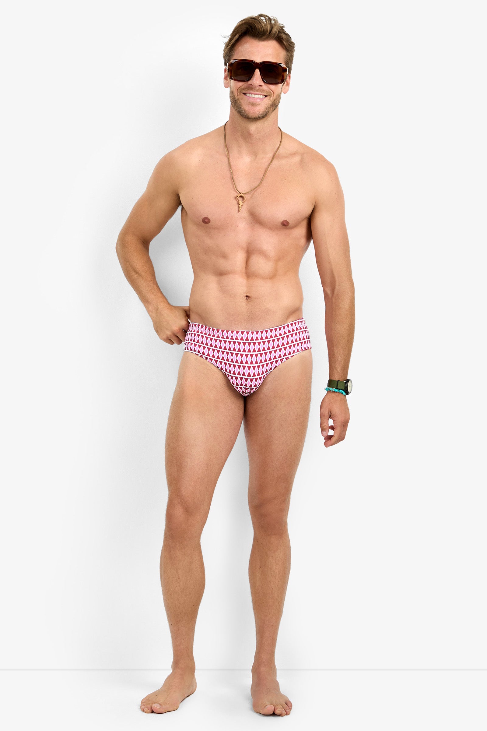 A smiling man models the BONDI SWIM BRIEF—red and patterned—paired with sunglasses, a necklace, and a wristwatch, standing barefoot against a plain white background.