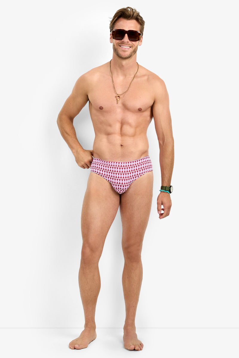 A smiling man models the BONDI SWIM BRIEF—red and patterned—paired with sunglasses, a necklace, and a wristwatch, standing barefoot against a plain white background.