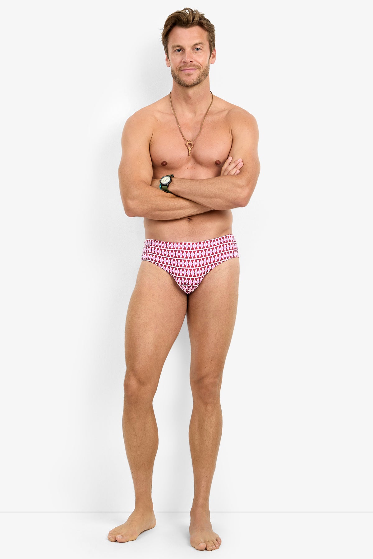 A man stands barefoot against a white background, wearing the BONDI SWIM BRIEF, a necklace, and a wristwatch. He has his arms crossed and smiles slightly.