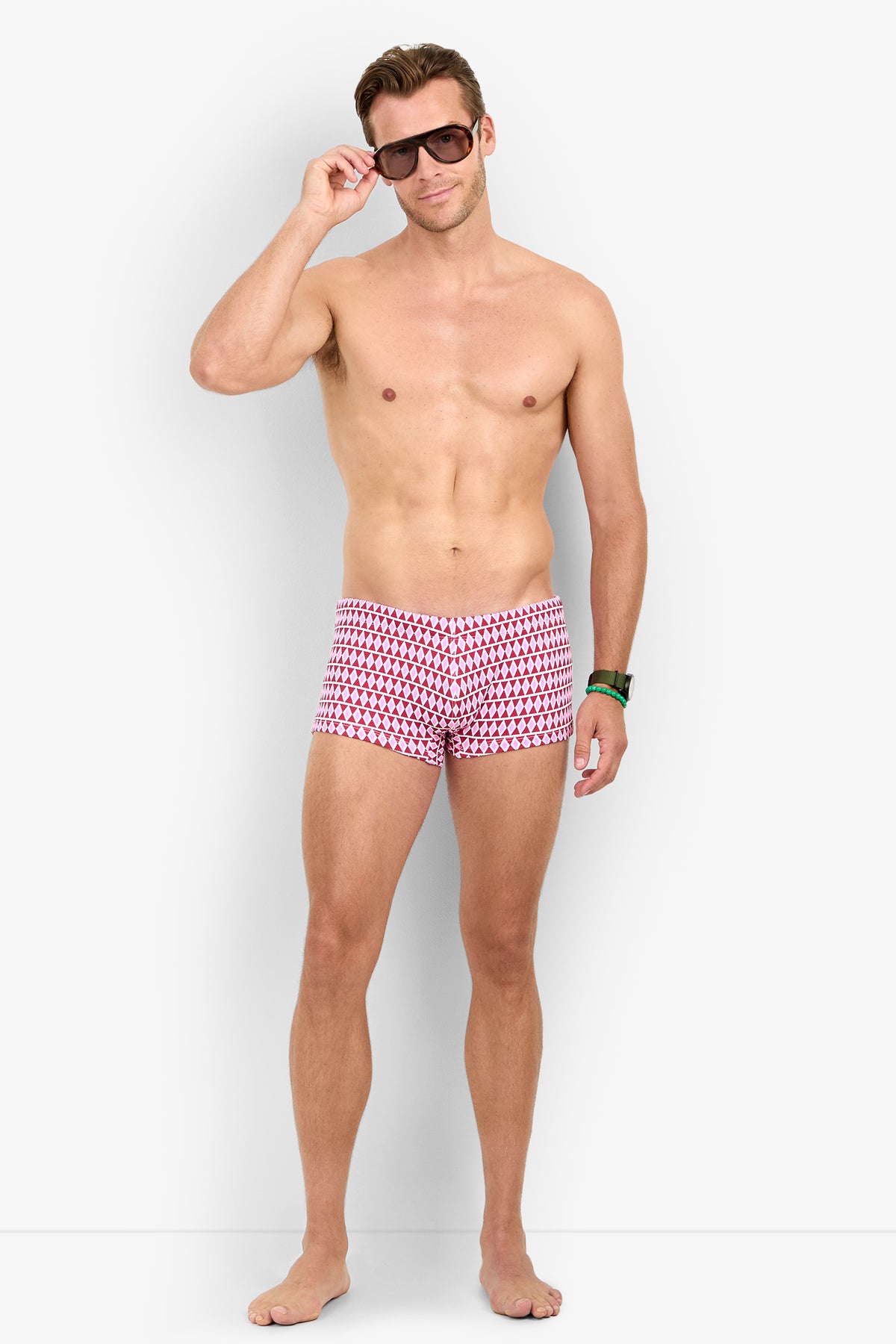 A shirtless man in MONTEGO BAY SWIM TRUNK and sunglasses stands barefoot against a plain white background, smiling slightly.