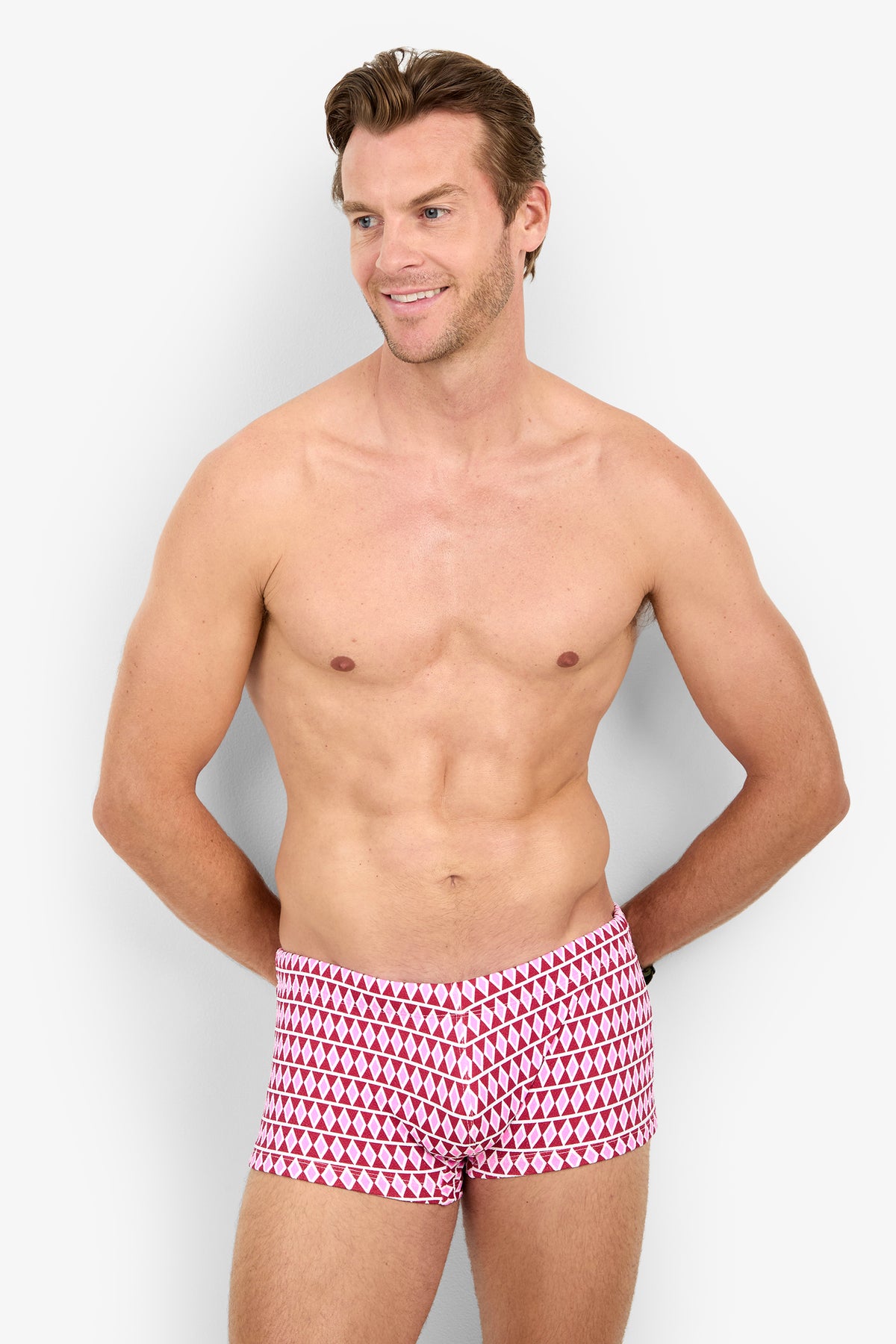 A smiling shirtless man stands with his hands behind his back, wearing the MONTEGO BAY SWIM TRUNK in a red and pink pattern against a plain white background.