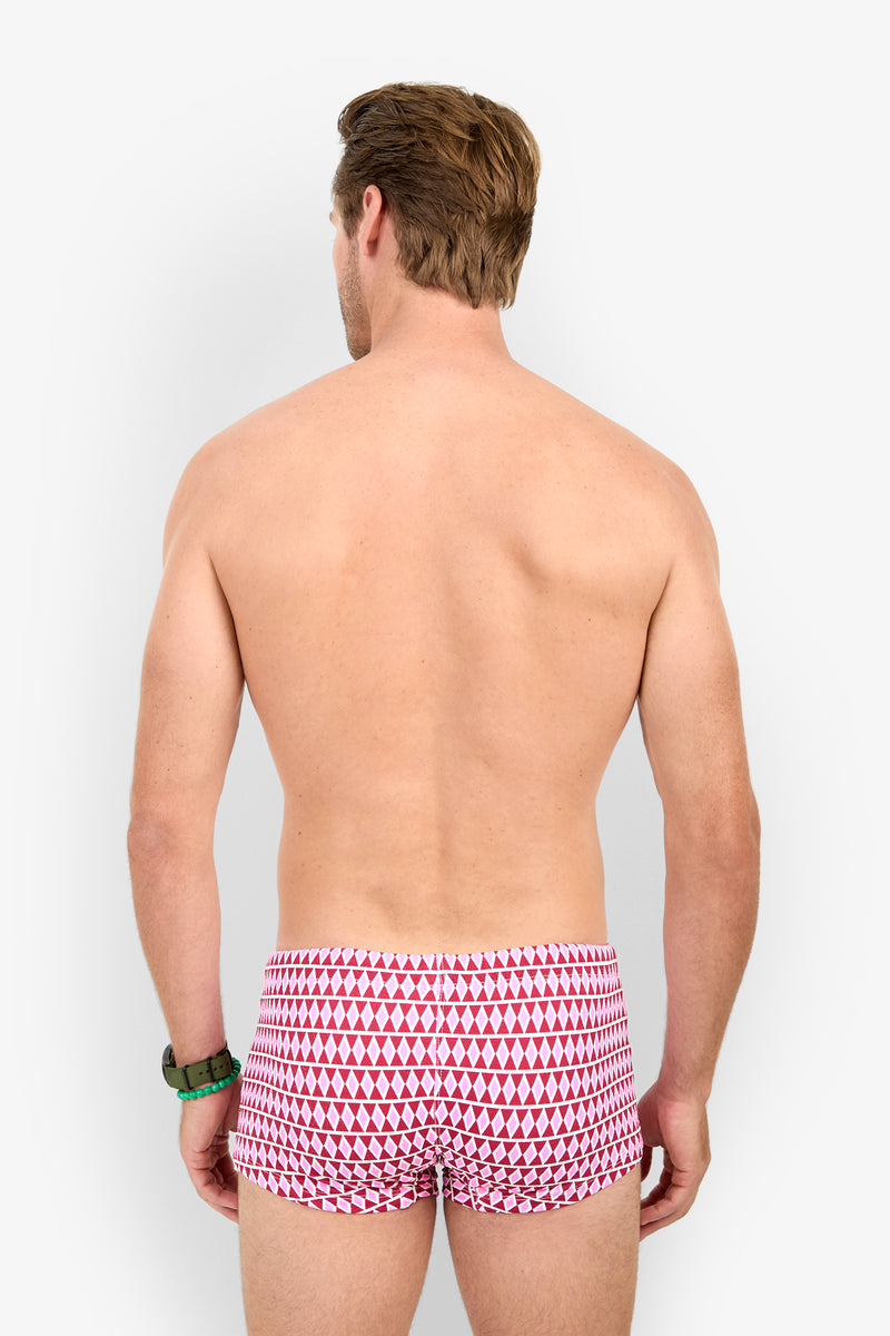 A shirtless man stands facing away, wearing MONTEGO BAY SWIM TRUNK in red and white pattern with a green wristwatch, set against a plain light background.