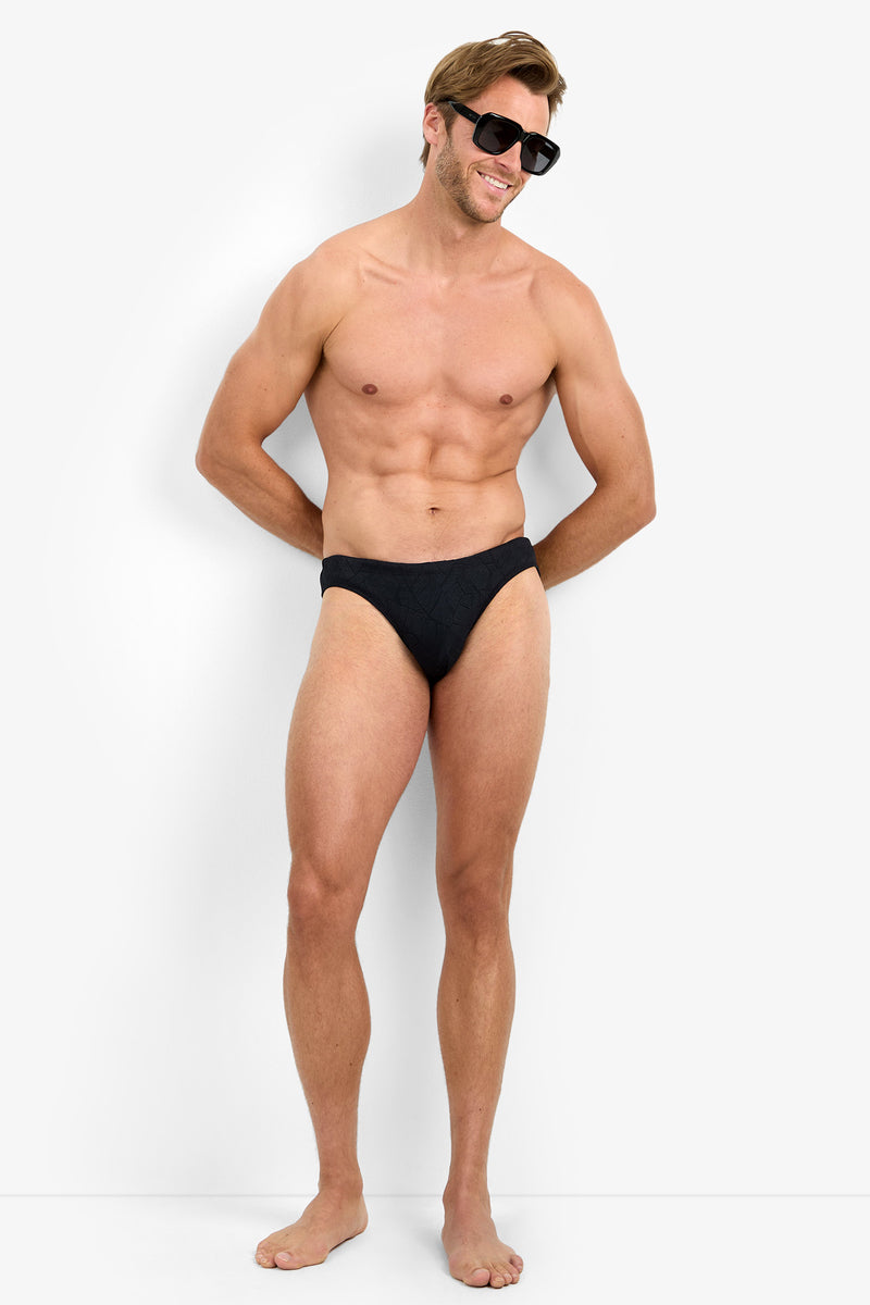 A smiling, muscular man stands barefoot against a plain light background, wearing LAGOS SWIM BRIEF and sunglasses with his hands behind his back.