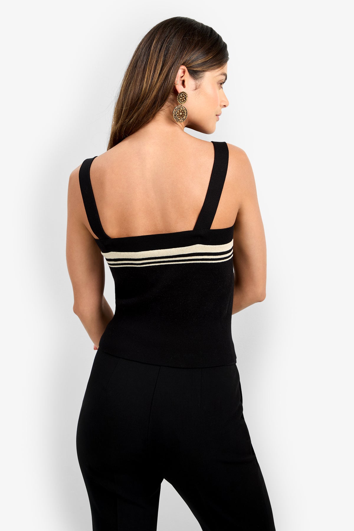 A woman with long brown hair, wearing large circular earrings and the CASADY CAMI—a black sleeveless top with two white stripes across the back—paired with black pants, stands against a plain light background, turned away from the camera.