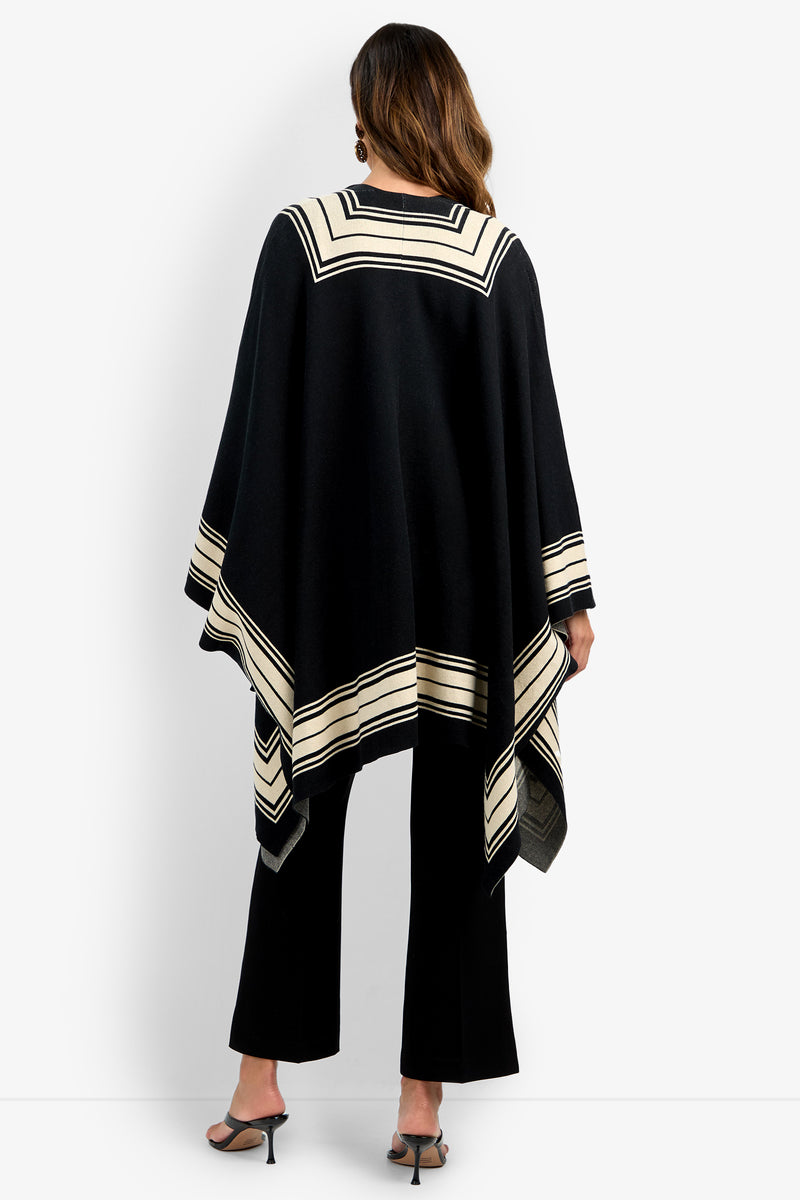 A woman with wavy brown hair models the VILLAS PONCHO, featuring black fabric with cream geometric stripe accents. She pairs it with black pants and heeled sandals, posing against a plain white background.