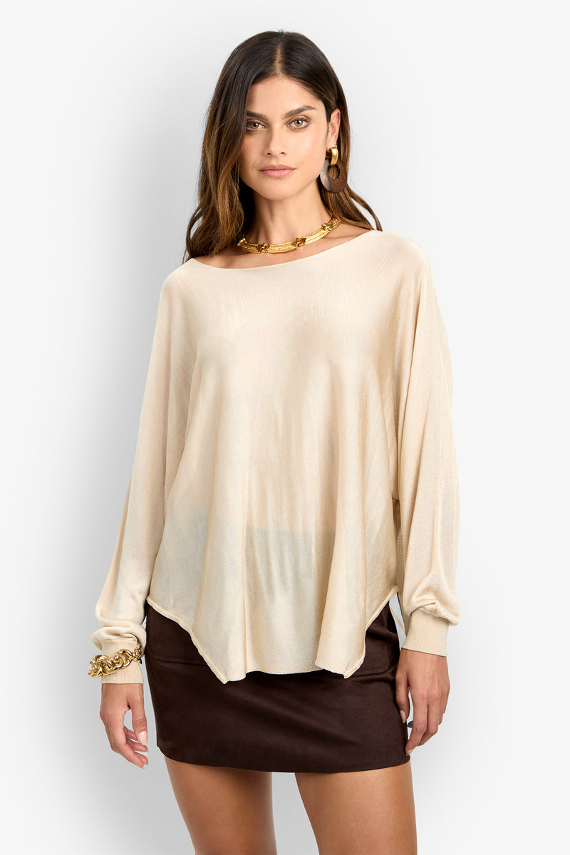 A woman wears the VINO PULLOVER, a loose long-sleeved beige top, with a short dark brown skirt, gold hoop earrings, a chunky gold necklace, and a gold bracelet while standing facing forward.