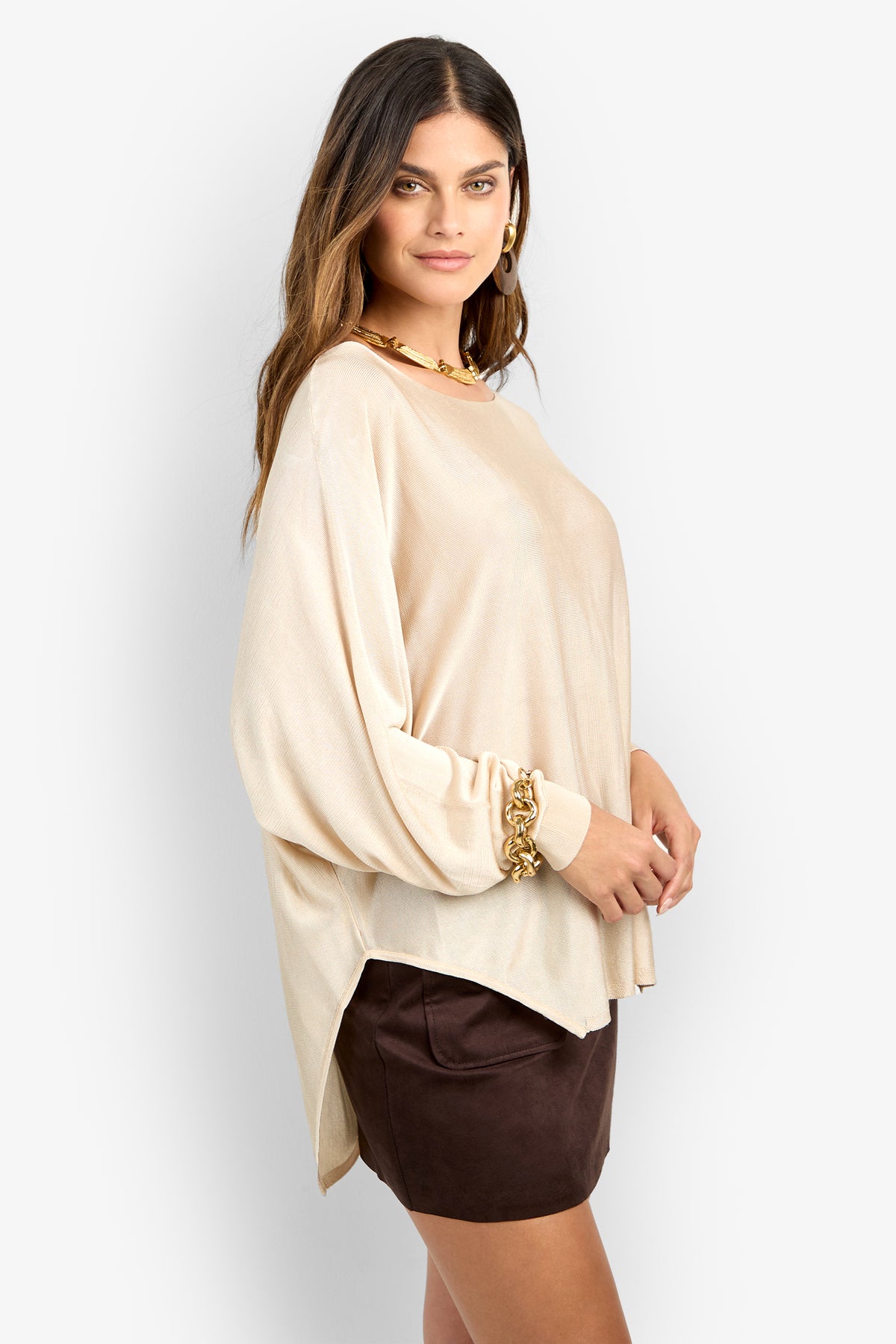 Woman standing sideways wearing the VINO PULLOVER, a loose beige long-sleeve top, paired with gold jewelry and a dark brown mini skirt, posing against a plain light background.