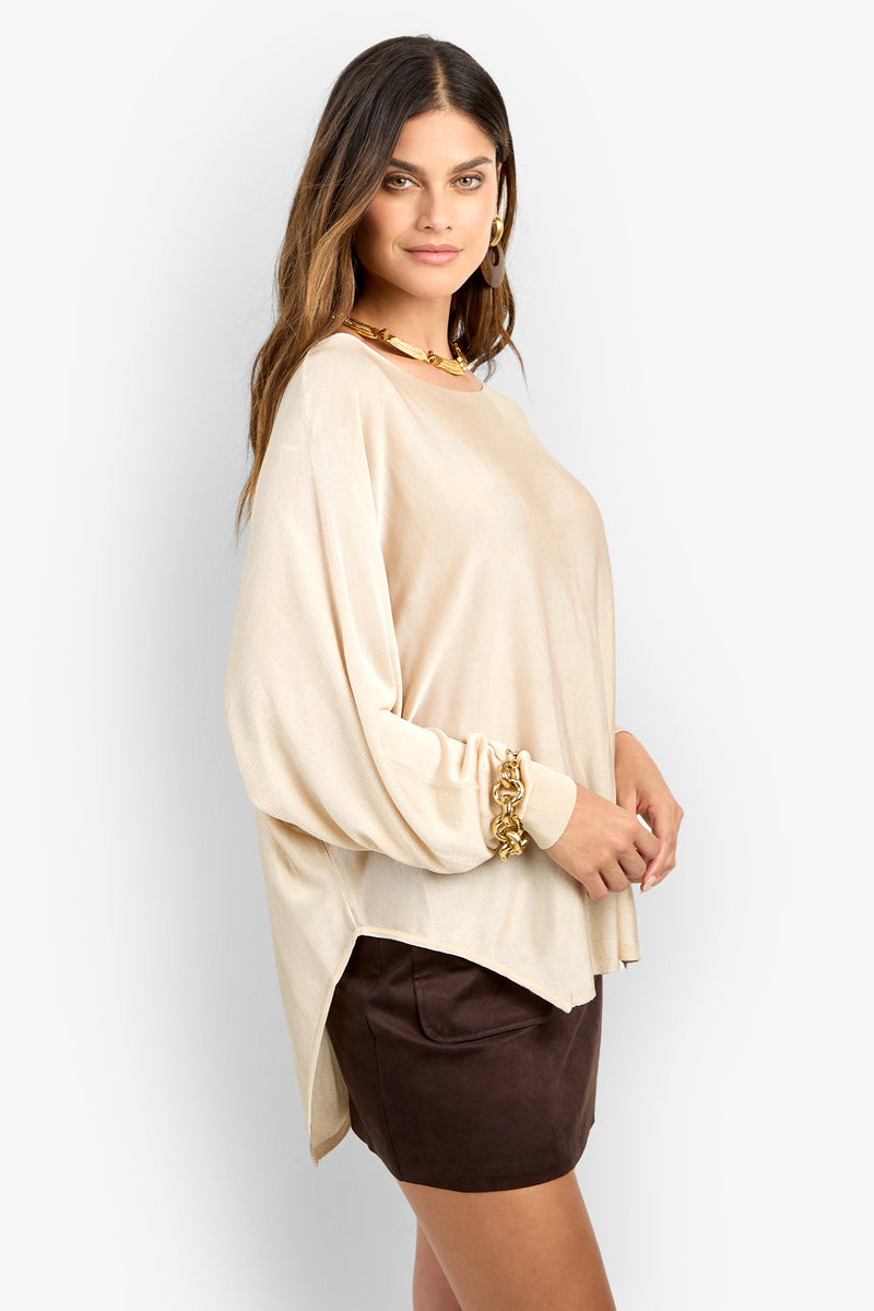 Woman standing sideways wearing the VINO PULLOVER, a loose beige long-sleeve top, paired with gold jewelry and a dark brown mini skirt, posing against a plain light background.