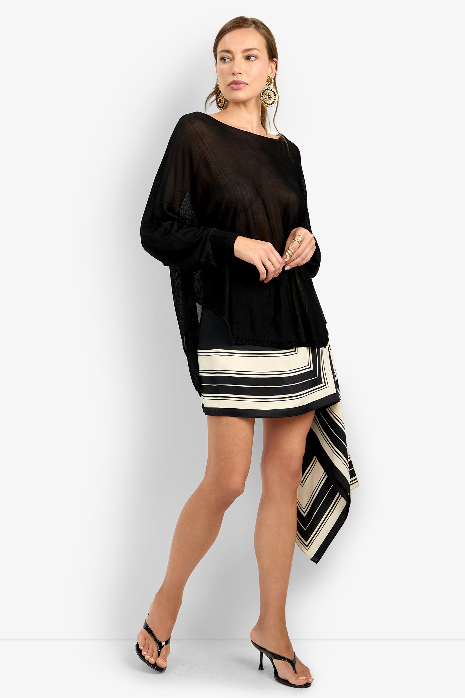 Woman wearing the VINO PULLOVER, a loose black long-sleeve top, paired with a black and white asymmetrical striped skirt, black heels, hoop earrings, and rings, standing against a plain white background.