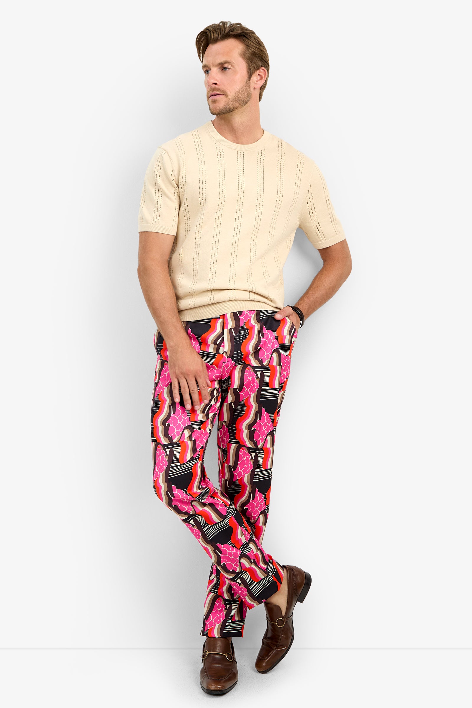 A man stands against a white background wearing the CARLO CREWNECK short-sleeve beige sweater, vibrant pink patterned pants, and brown loafers, with one hand in his pocket and the other resting by his side.