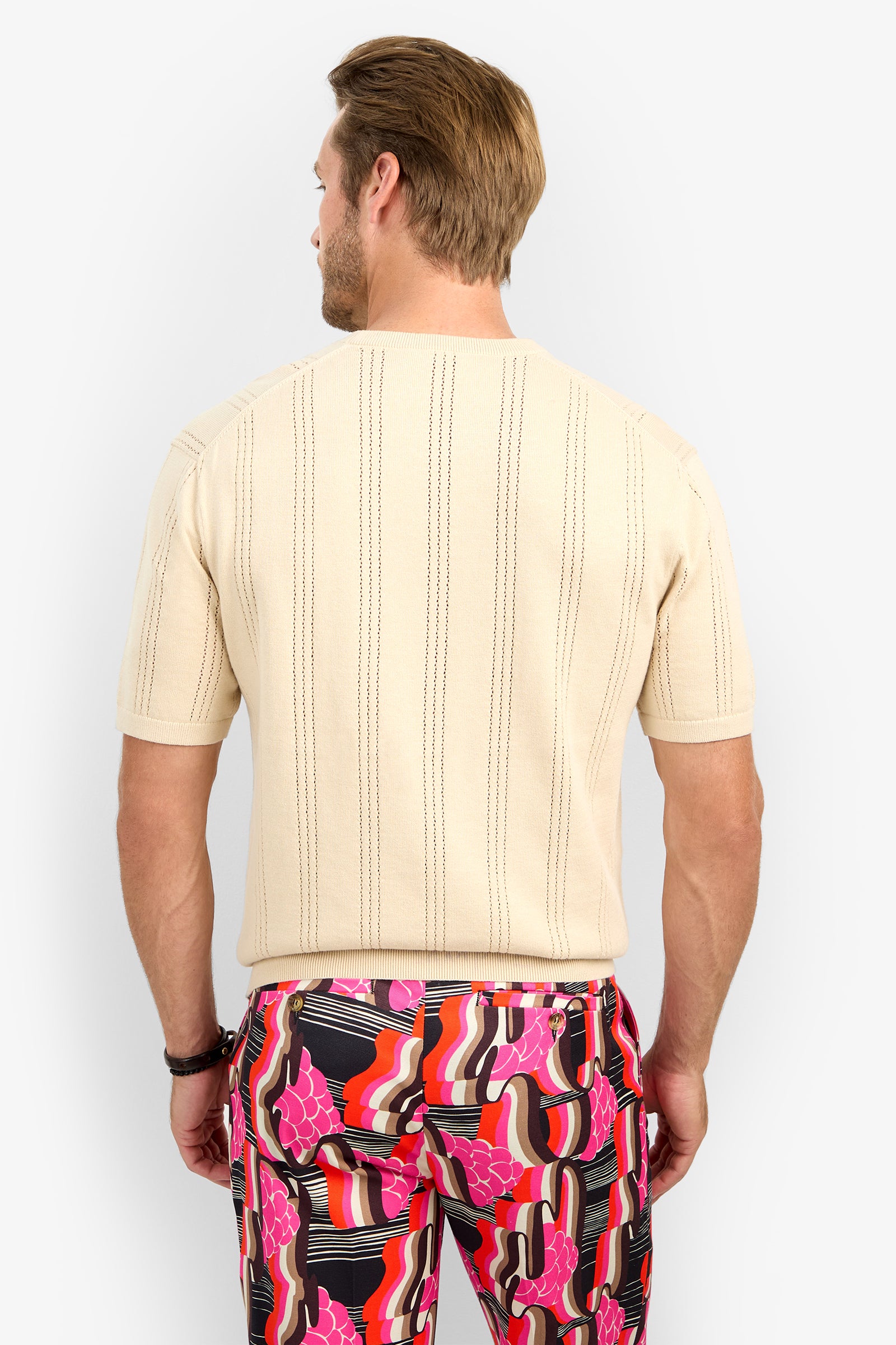 A man is seen from behind wearing the CARLO CREWNECK—a short-sleeved, beige textured shirt—paired with bold pants featuring a pink, red, black, and white abstract pattern.