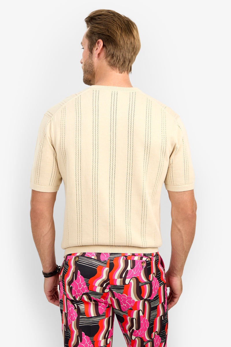 A man is seen from behind wearing the CARLO CREWNECK—a short-sleeved, beige textured shirt—paired with bold pants featuring a pink, red, black, and white abstract pattern.