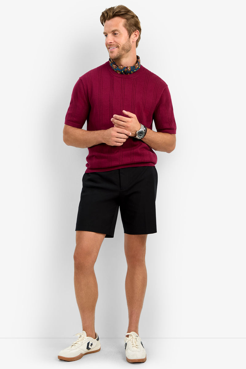 A man stands against a white background wearing the CARLO CREWNECK short-sleeve maroon sweater, a floral scarf, black shorts, white sneakers with black accents, and a wristwatch, looking off to the side and smiling.