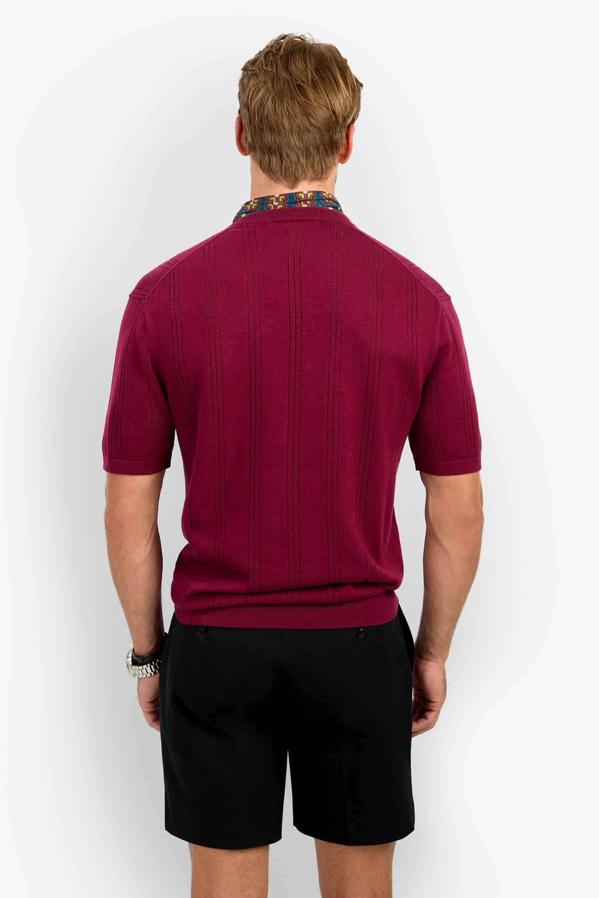 A man with light brown hair and a watch on his left wrist stands facing away, wearing the CARLO CREWNECK short-sleeved textured burgundy shirt and black shorts against a plain white background.