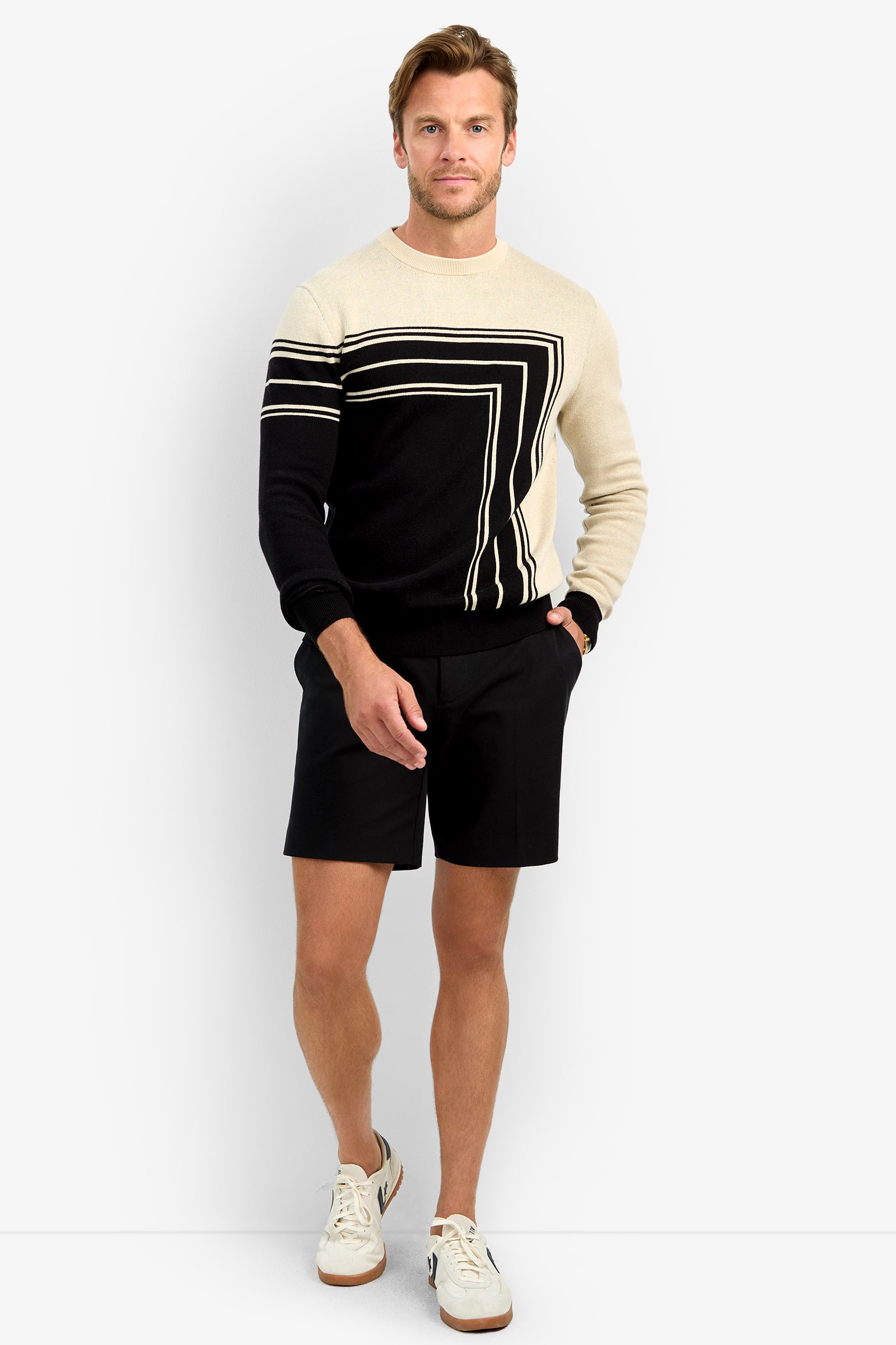 A man stands against a plain white background wearing the RAMOS STRIPE CREWNECK, black shorts, and white sneakers with brown soles. His left hand is in his pocket, and he smiles slightly.