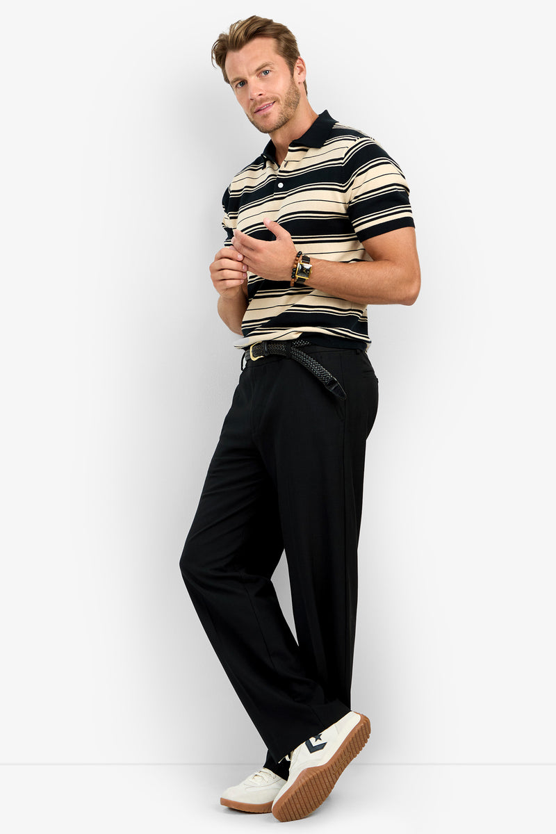 A man poses against a plain background wearing the EDWARD STRIPE POLO, black belted trousers, and white sneakers. He holds sunglasses and looks at the camera with a relaxed expression.