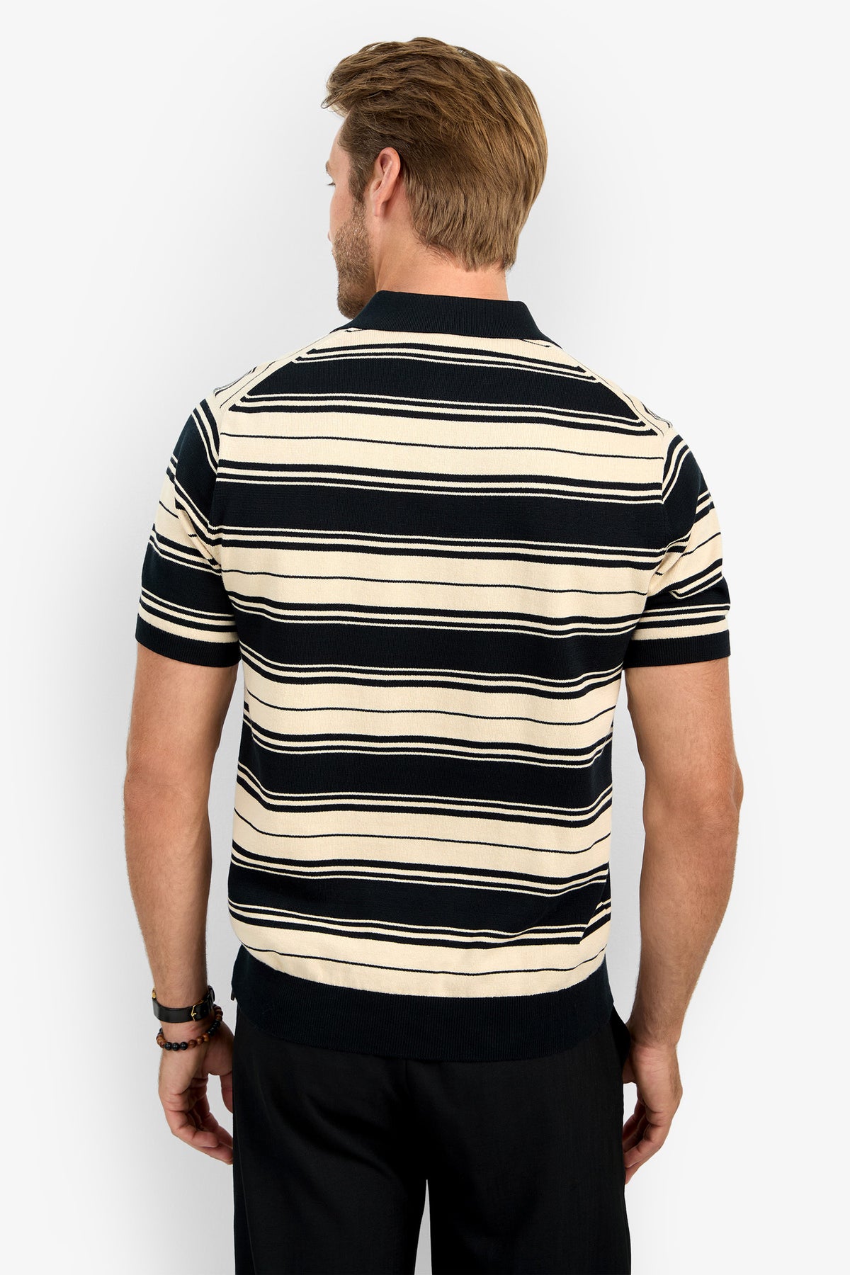 A man with light brown hair is seen from the back wearing the EDWARD STRIPE POLO, a short-sleeve collared knit shirt with black and beige horizontal stripes and black trim, paired with black pants.