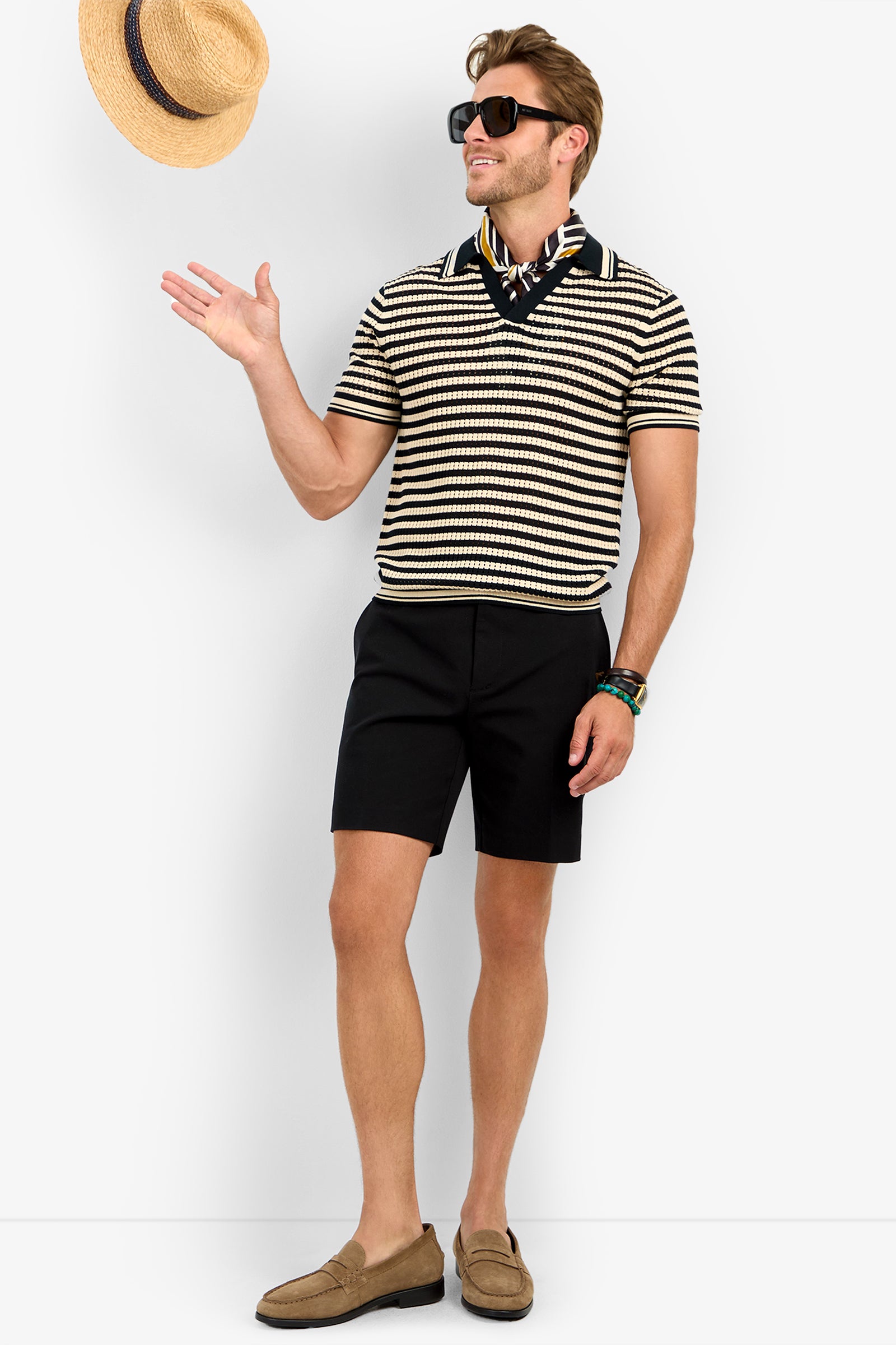 A man wearing sunglasses, the ALLEN MESH POLO, black shorts, loafers, and a patterned neck scarf smiles as he tosses a straw hat in the air.