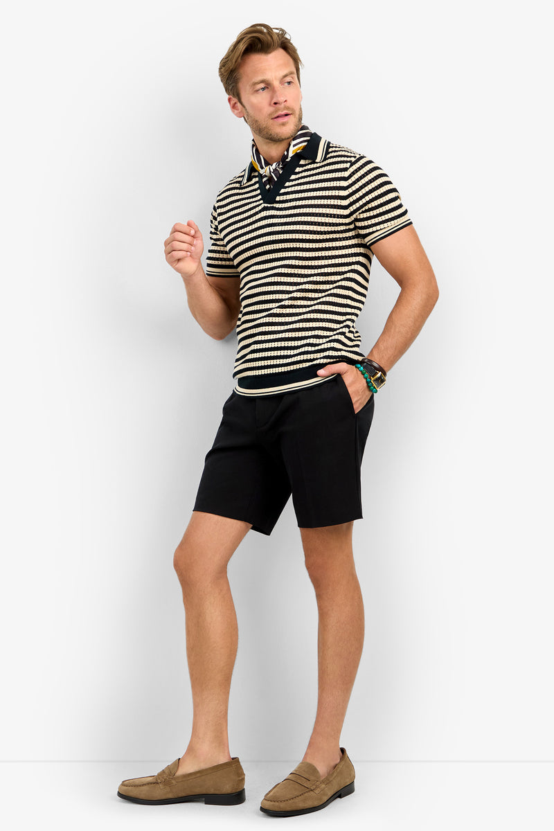 A man poses against a white background wearing the ALLEN MESH POLO, black shorts, a black neck scarf, a watch, and tan loafers. He stands with one hand raised and the other in his pocket.