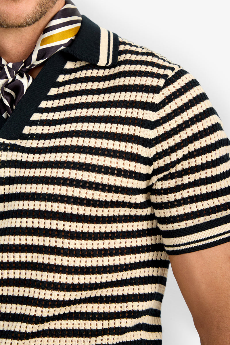 A person in the ALLEN MESH POLO, a short-sleeve striped knit shirt in cream, black, and brown with a wide collar, paired with a black, white, and yellow patterned scarf around their neck. Only their lower face, neck, and upper torso are shown.