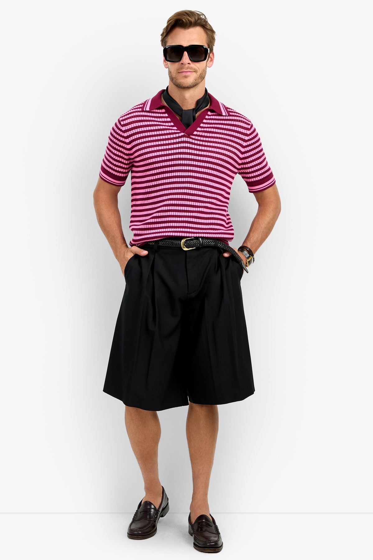 A man wearing sunglasses, an ALLEN MESH POLO in pink and white stripes, black long shorts, a black belt, brown loafers, and a watch stands with hands in his pockets against a plain white background.