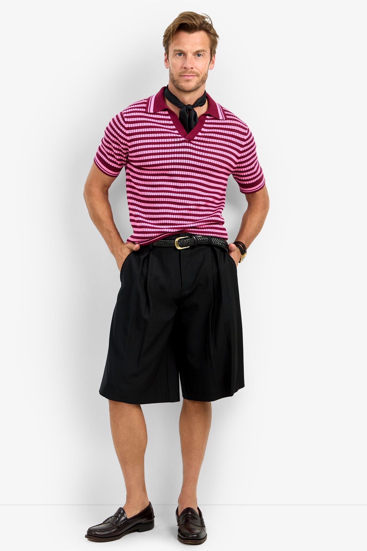 A man stands against a plain background wearing the ALLEN MESH POLO in pink and white stripes, a black scarf, black pleated knee-length shorts with a black belt, and brown loafers. One hand is in his pocket as he smiles slightly.