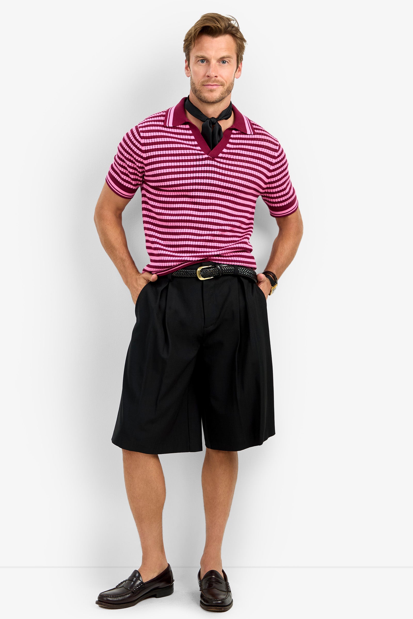A man stands against a plain background wearing the ALLEN MESH POLO in pink and white stripes, a black scarf, black pleated knee-length shorts with a black belt, and brown loafers. One hand is in his pocket as he smiles slightly.