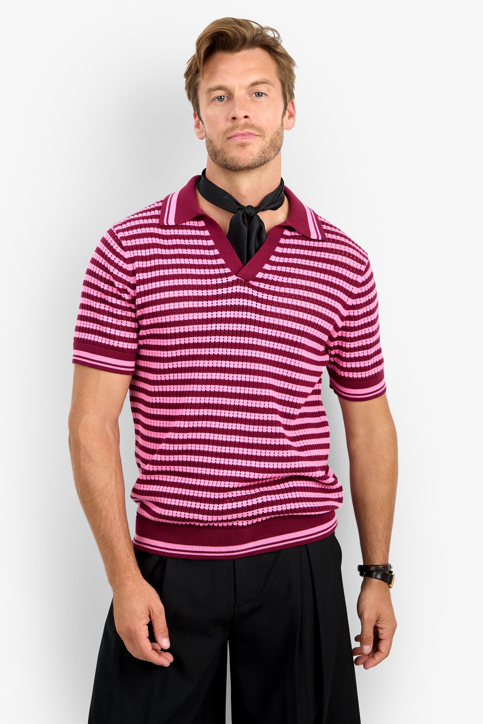 A man with light brown hair and a short beard wears the ALLEN MESH POLO, which is pink and white striped with short sleeves, paired with a black scarf, black pants, and a watch, standing against a plain white background.