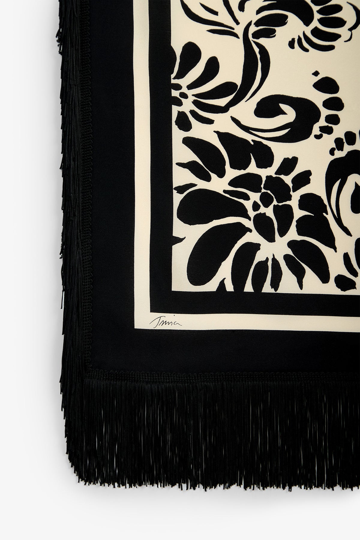 A cream-colored Tivoli Fringe Scarf featuring black floral patterns, a black border, black fringe along the edge, and a small signature near the corner.