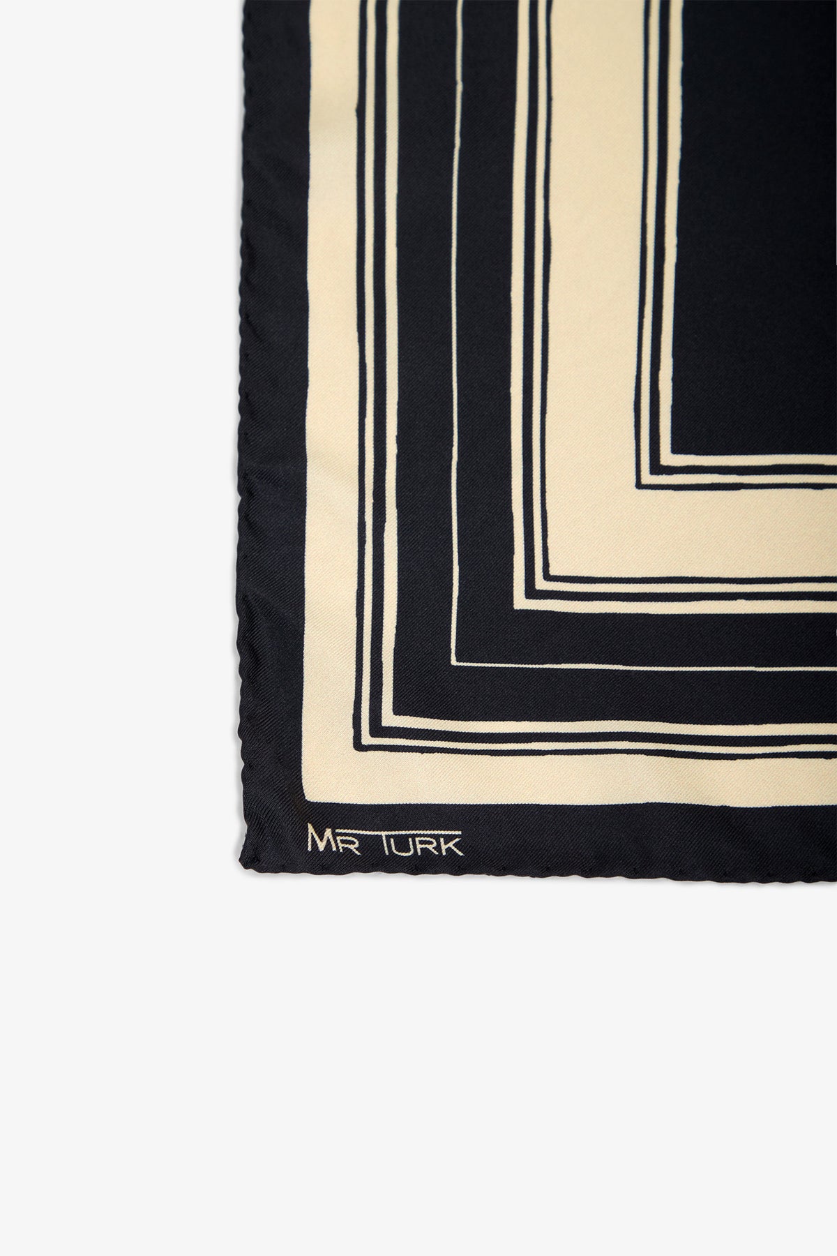 Corner of a black and cream patterned Pocket Square featuring geometric lines, with the words MR TURK stitched in white in the lower left corner.