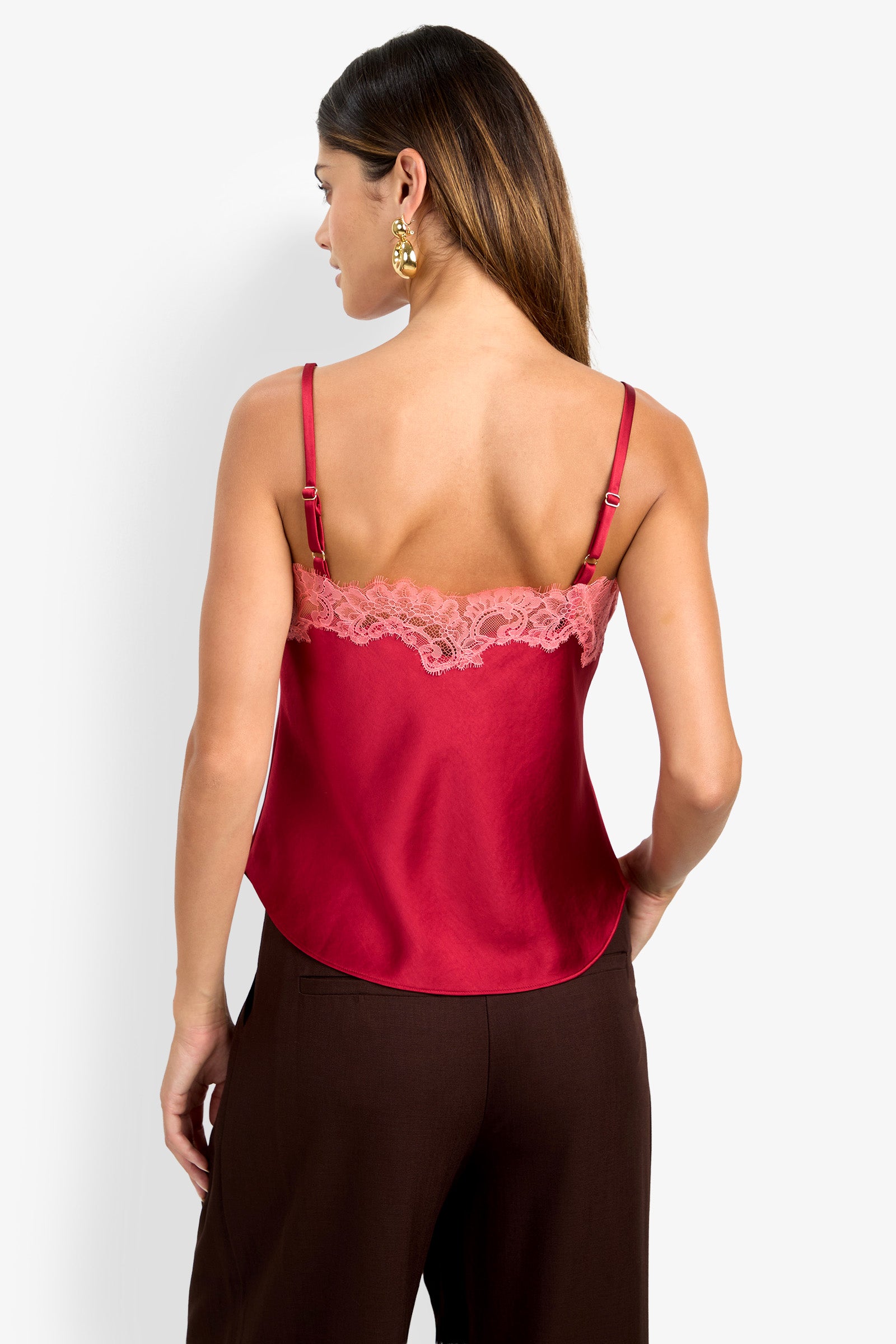 A woman with long, straight hair and gold earrings stands facing away against a plain white background, wearing the Amoira Top—a red satin camisole with pink lace trim and adjustable straps—paired with brown pants.