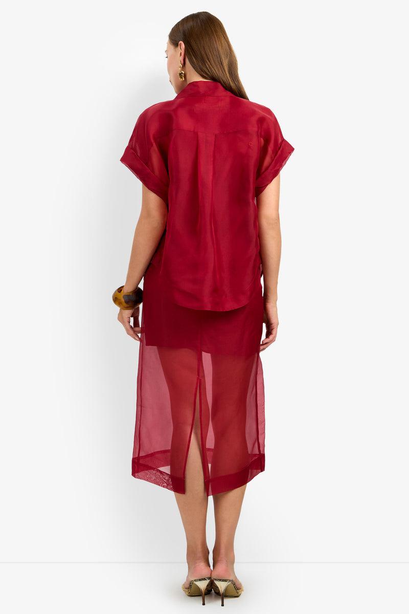 A woman stands against a white background, facing away and wearing the COSTANZO TOP—a sheer red short-sleeve shirt—layered over a matching red dress or slip, paired with heels and hoop earrings.