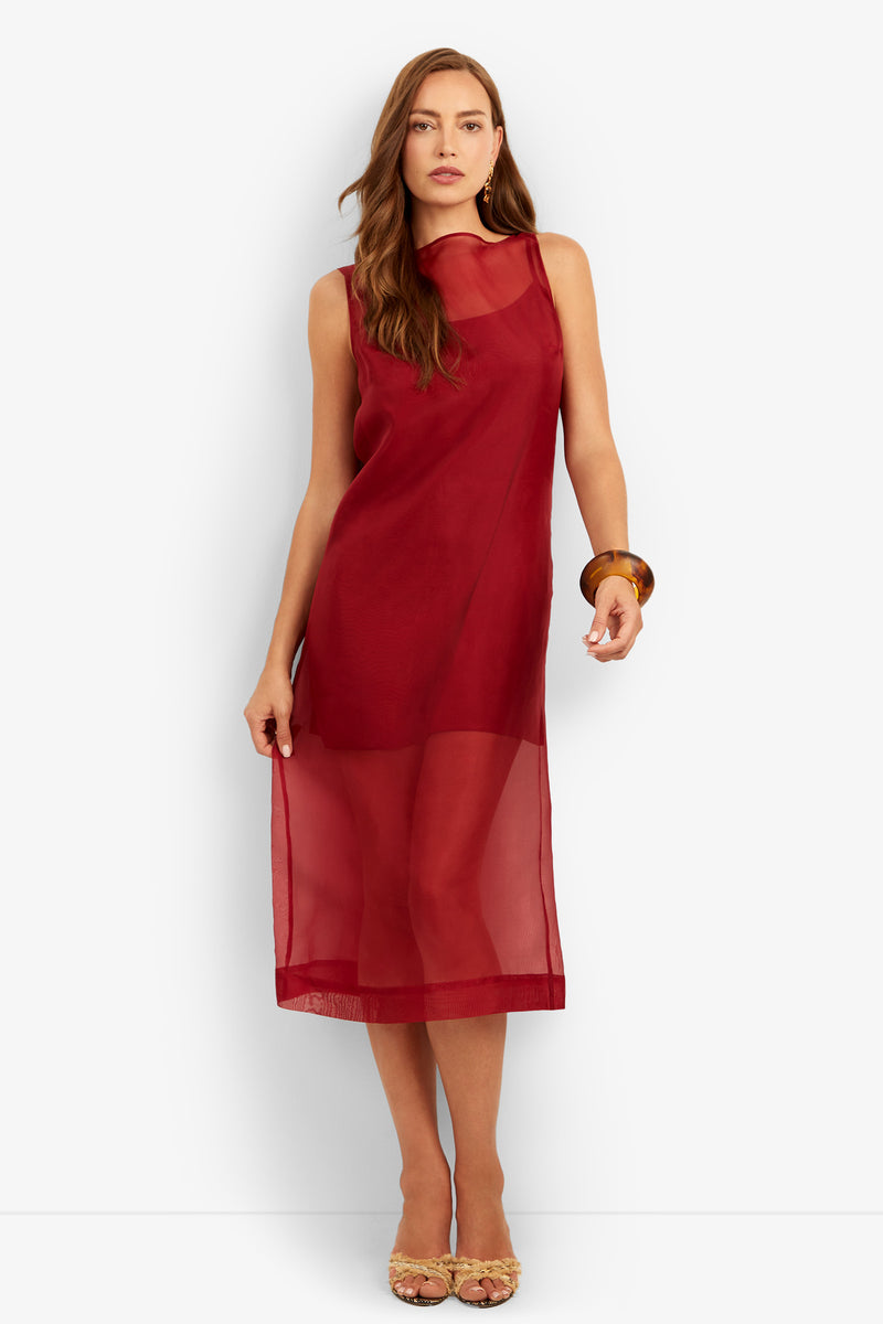 A woman stands against a plain white background wearing the Roselle Dress—a sleeveless, sheer red midi dress with a high neckline—paired with nude heels and a chunky bracelet.