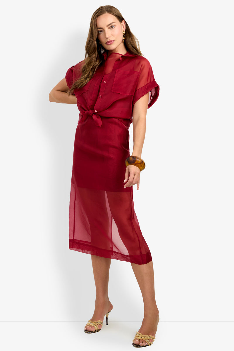 Woman wearing the COSTANZO TOP, a sheer red button-up style with short sleeves and a tied waist, styled as a dress with a midi-length skirt. She accessorizes with a brown bangle and beige heeled sandals, standing against a plain white background.