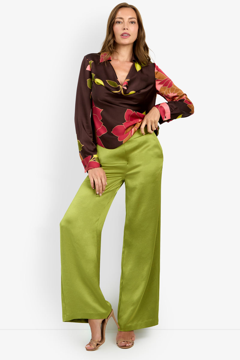 A woman poses in the Mistic Top, a brown floral blouse with pink and yellow accents, paired with high-waisted, wide-leg green satin pants and beige open-toe heels, standing against a plain white background.