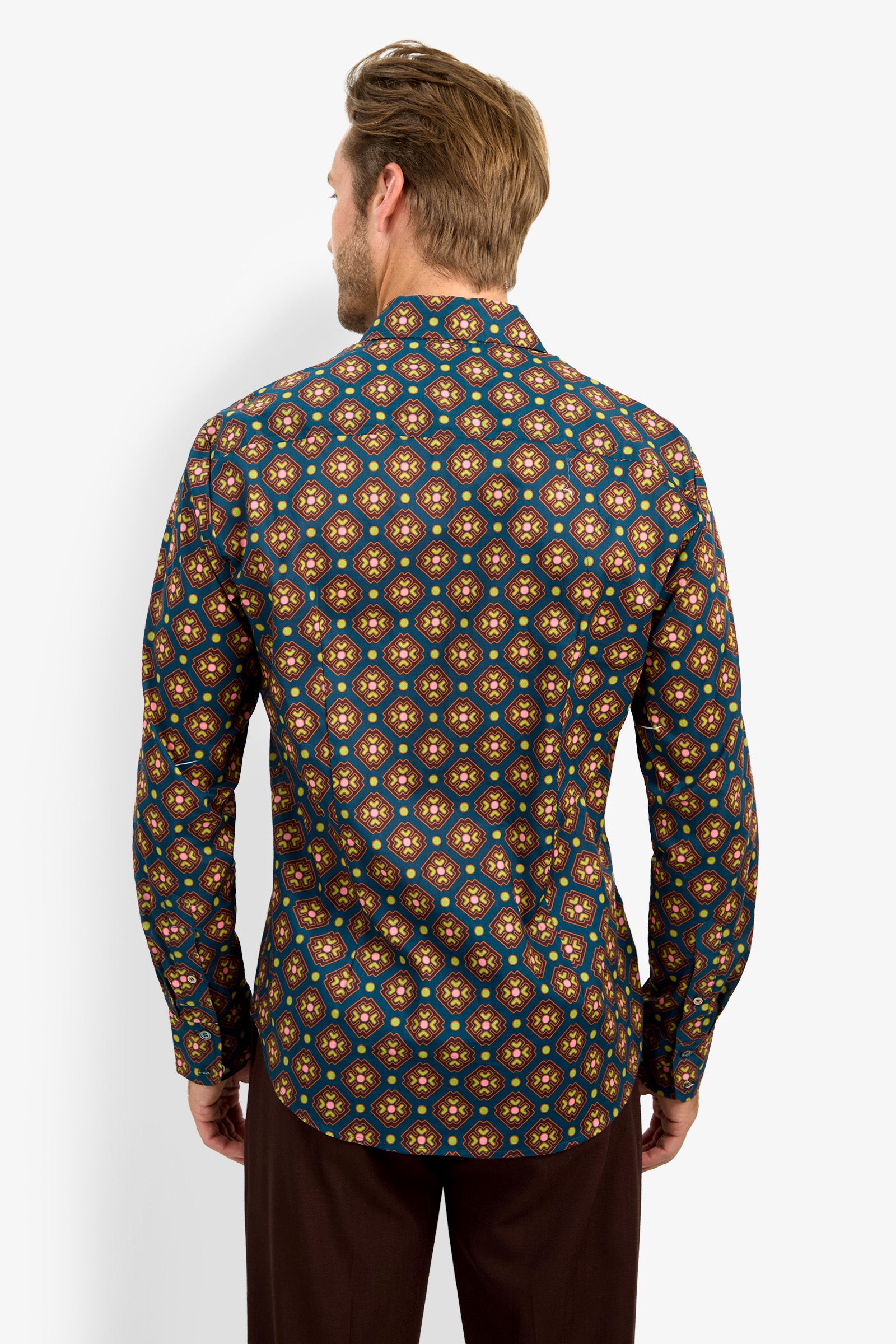 A man with light brown hair stands facing away, wearing the Aaron Shirt—a long-sleeved top featuring a vibrant geometric pattern in yellow, orange, and blue tones—paired with dark pants against a plain background.