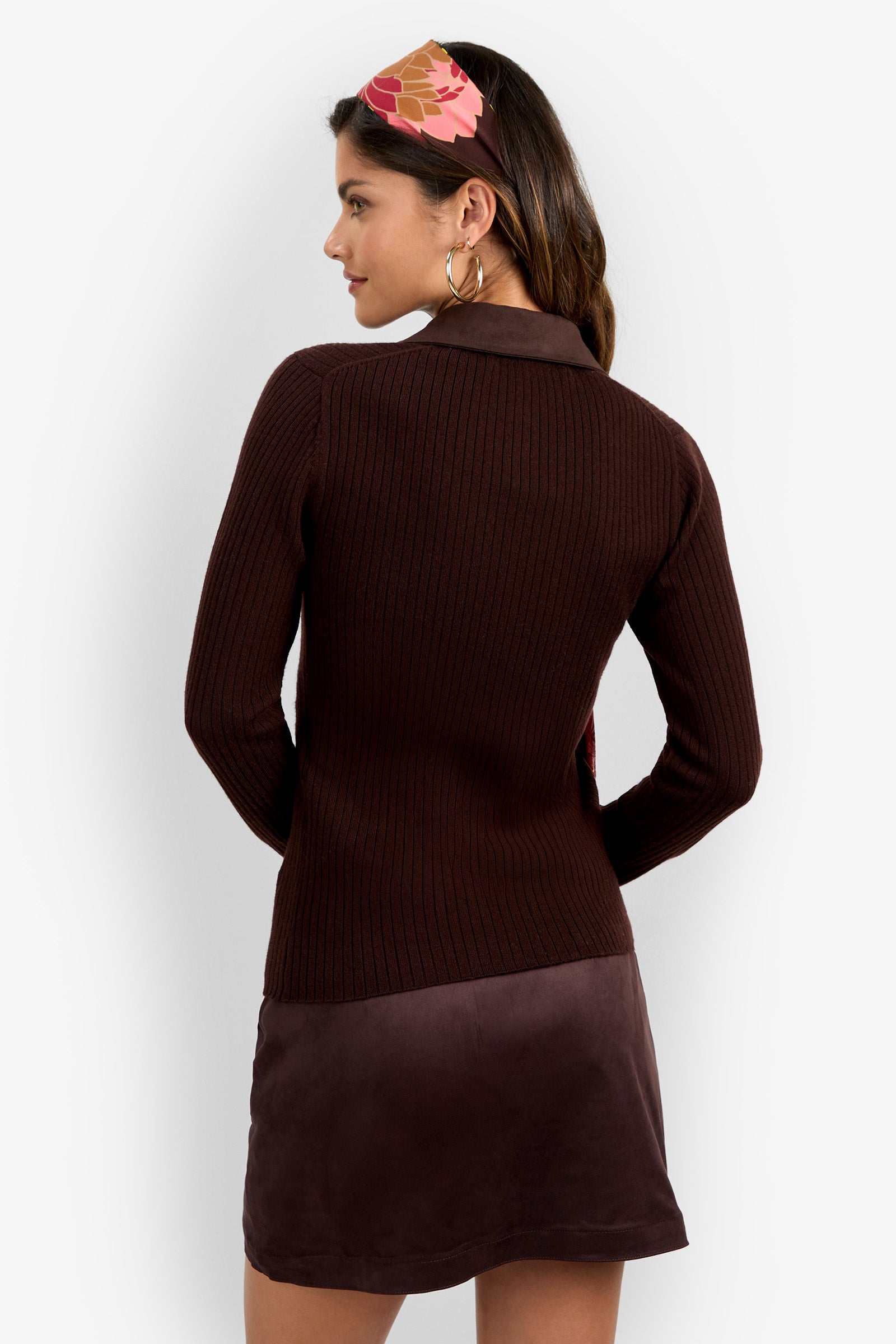 A woman with long brown hair, facing away, wears the Rita Long Sleeve Polo—a ribbed, dark brown top—paired with a mauve skirt. She accessorizes with large hoop earrings and a pink and tan geometric hairband.
