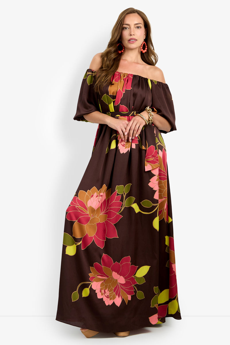 A woman models the Voglia Dress—an off-the-shoulder brown maxi dress featuring bold pink and orange floral patterns. She accessorizes with red earrings and bracelets, standing against a plain white background.